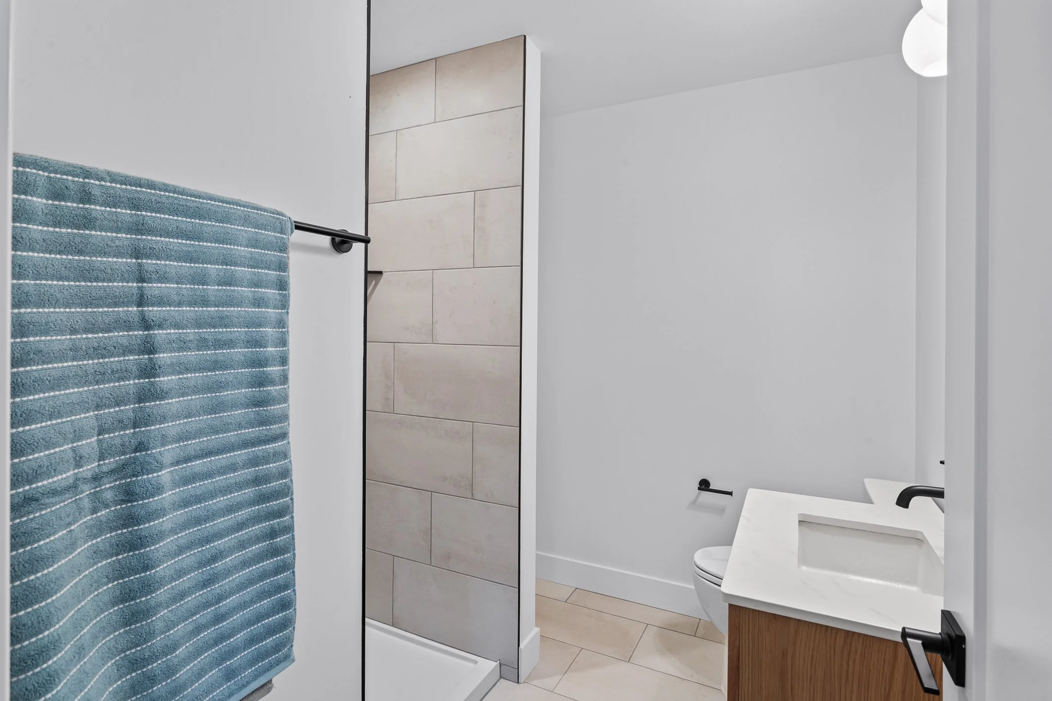 Modern bathroom with white walls, beige tiled floor, wooden vanity with white sink, black faucet, and a toilet. A blue towel hangs on a black towel bar.