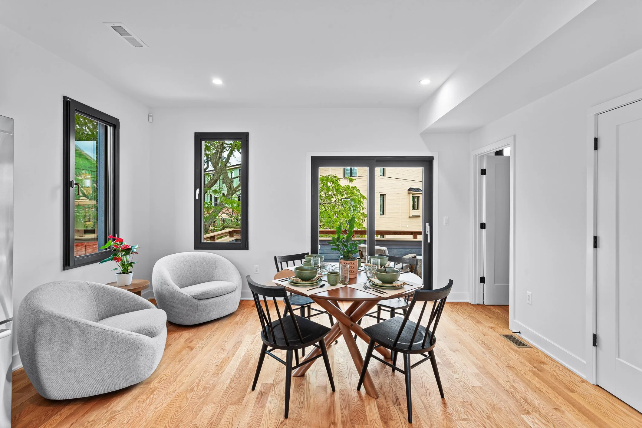 Modern dining room with a round wooden table set with dishes, surrounded by five black chairs, and two white armchairs by windows, with a door leading to an outdoor balcony with greenery.