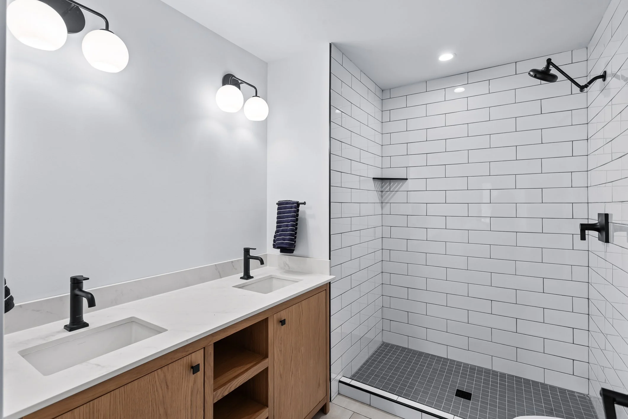 Modern bathroom featuring dual sinks with black fixtures, a large mirror, wall-mounted lighting, a towel rack, and a walk-in shower with white subway tiles, black fixtures, a corner shelf, and gray floor tiles.