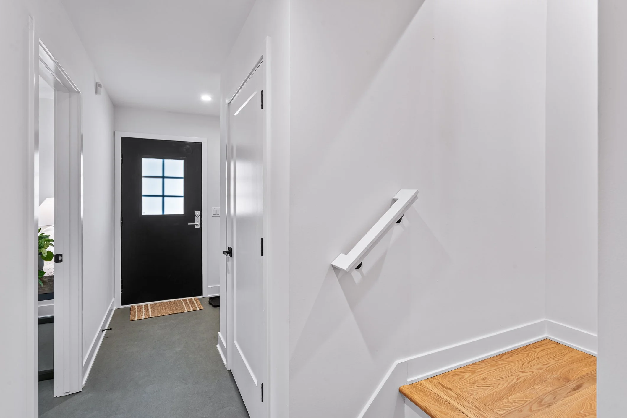 Interior view of a modern home hallway with white walls, a black front door with a small window, grey carpet, and white doors leading to rooms, with a wooden staircase landing and a small mat by the door.