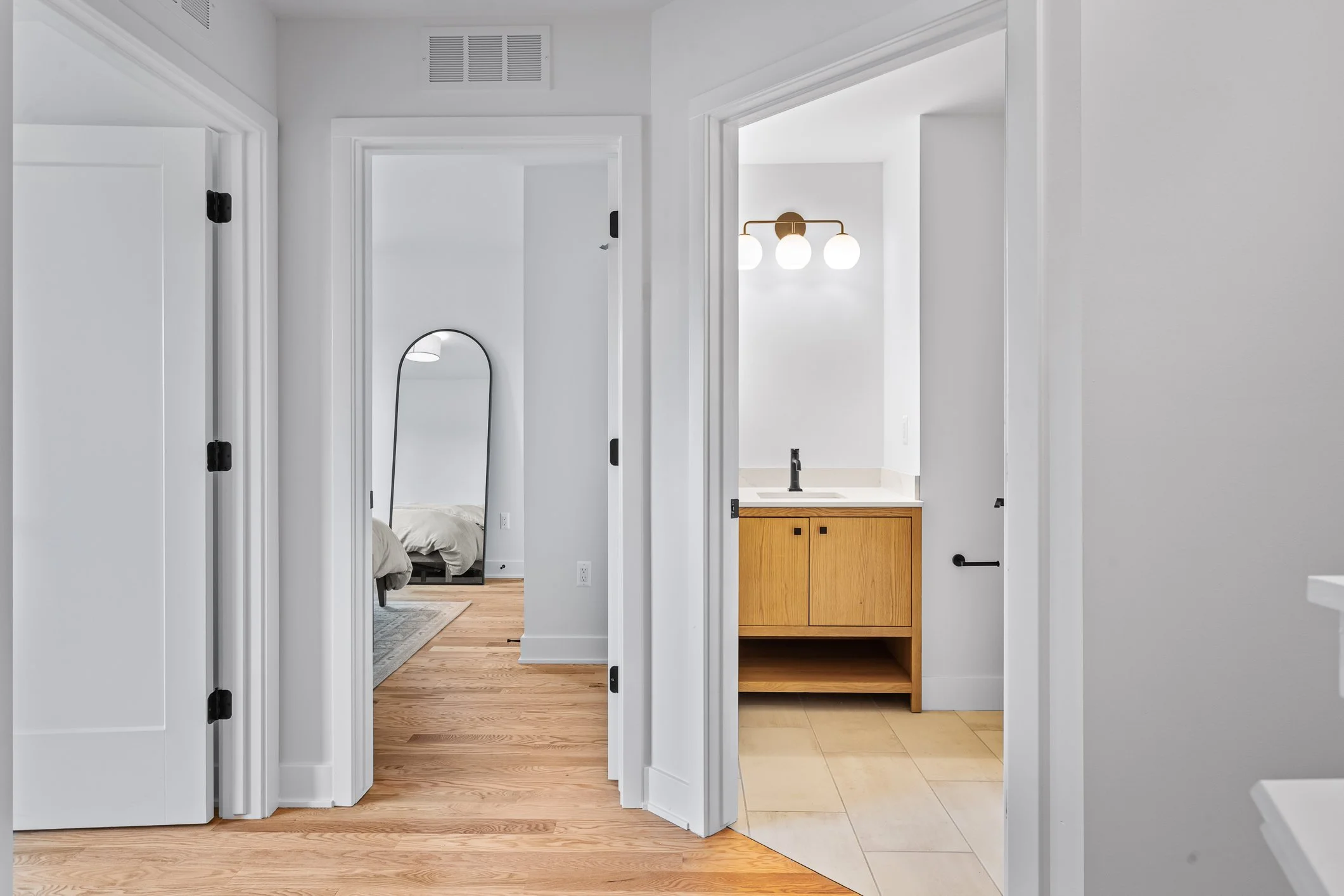 View of an interior hallway showing rooms with white walls, hardwood and tile flooring, a mirror and a bedroom in the background, and a bathroom with a wood cabinet and vanity lighting.