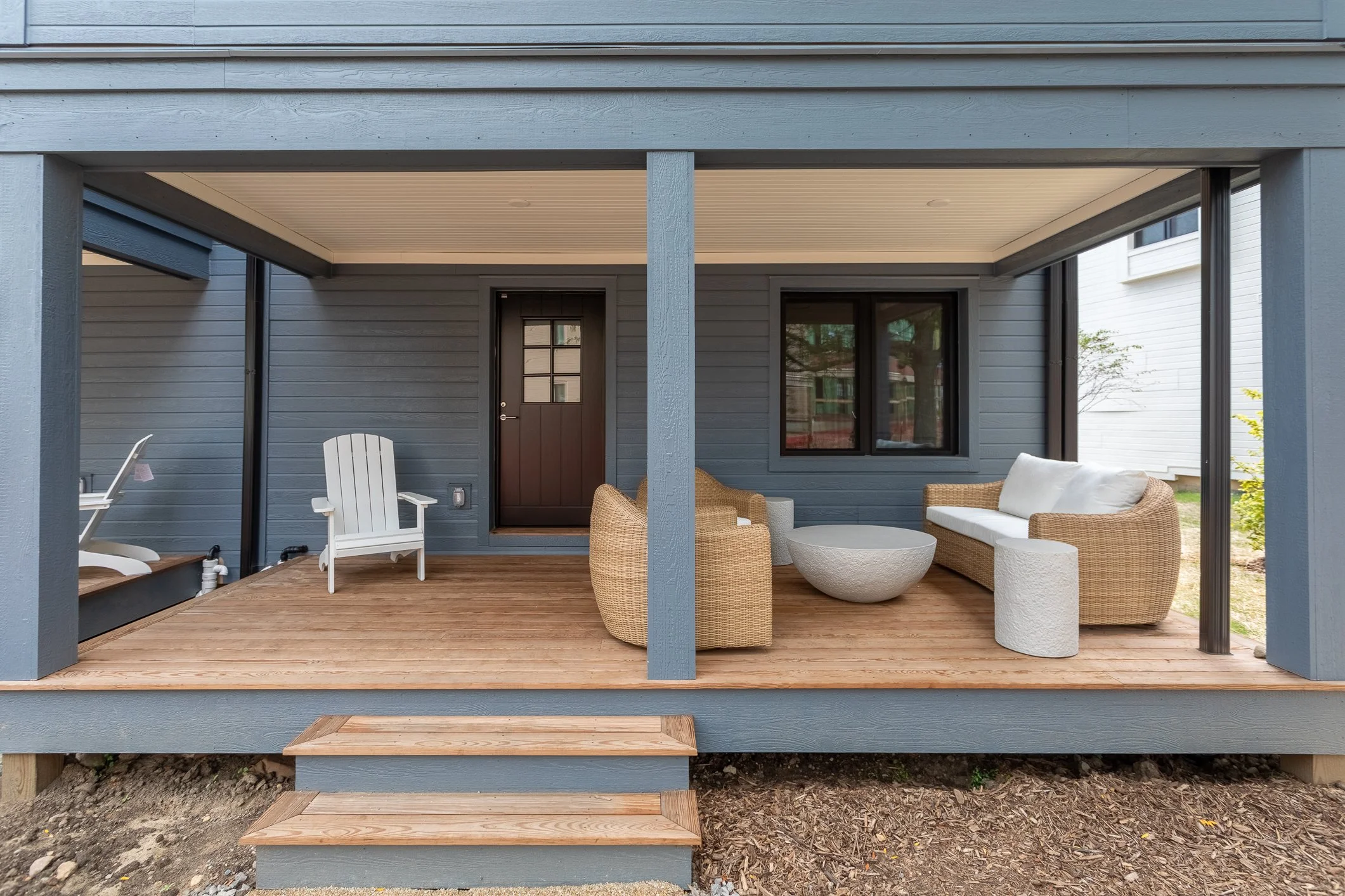 Covered front porch with wooden flooring, outdoor furniture including an Adirondack chair, wicker sofa with cushions, wicker armchairs, and a round stone coffee table. White and black exterior of a house, with a front door and window visible.