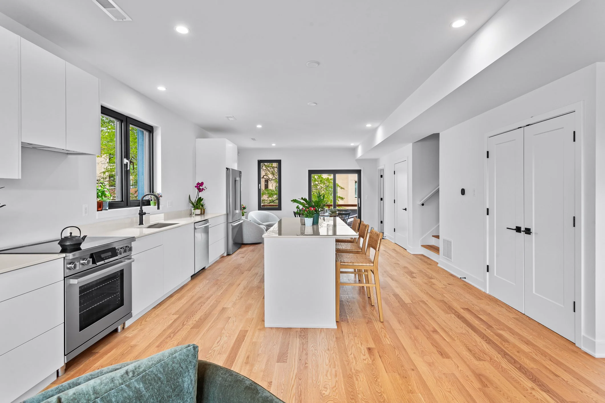 Modern kitchen with white cabinets, stainless steel appliances, and hardwood floors, featuring an island with chairs and a seating area with gray chairs and a table near windows with views of green trees.