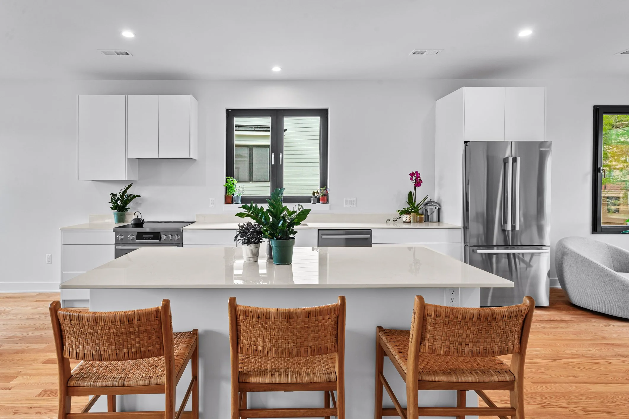 Modern kitchen with white cabinets, stainless steel refrigerator, a large central island with wooden chairs, and various potted plants on the counter and window sill.
