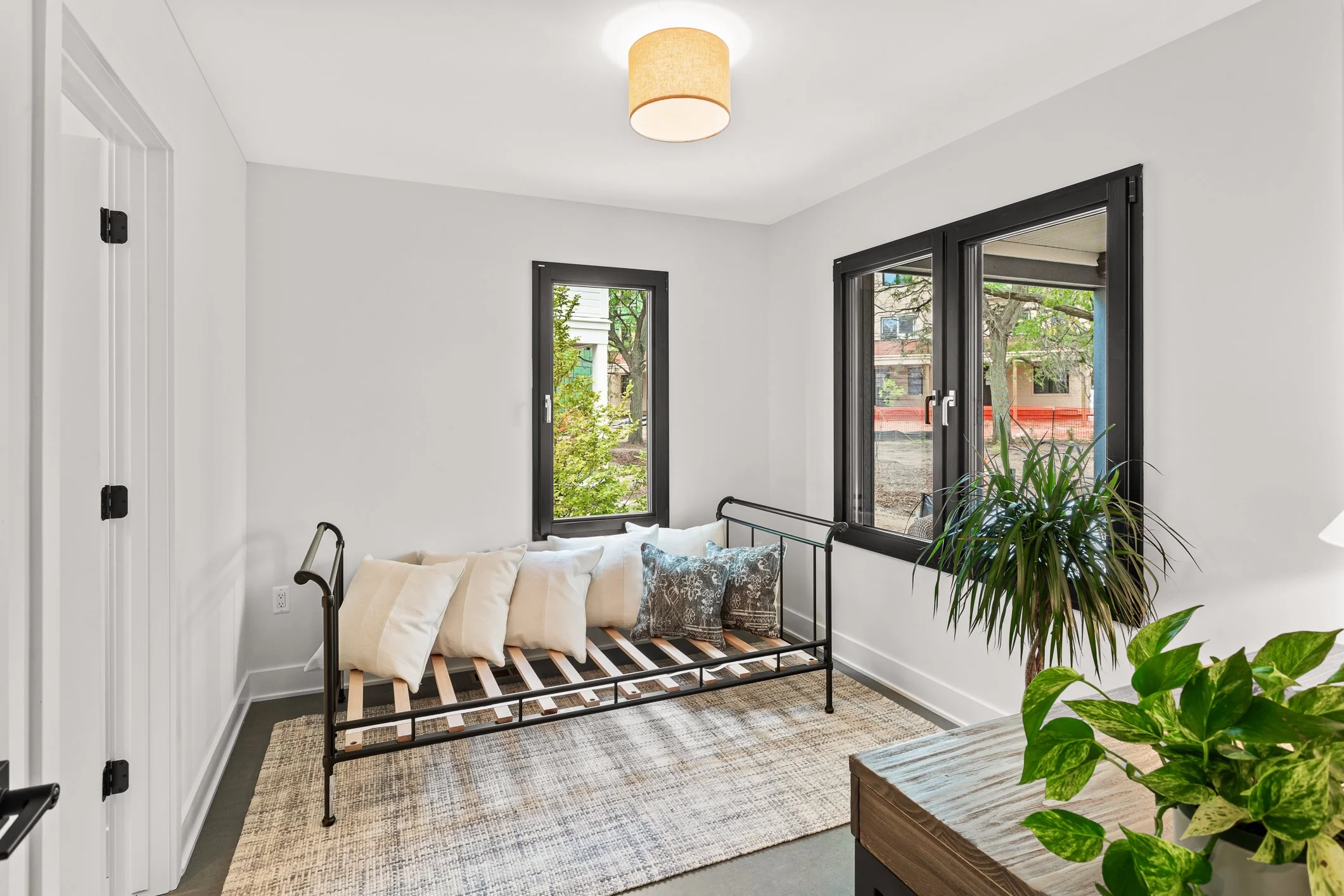 A bright room with white walls, black-framed windows, and a light-colored ceiling. There is a black metal daybed with wooden slats, topped with several white and patterned pillows, next to a beige and brown woven area rug. A wooden table with a green