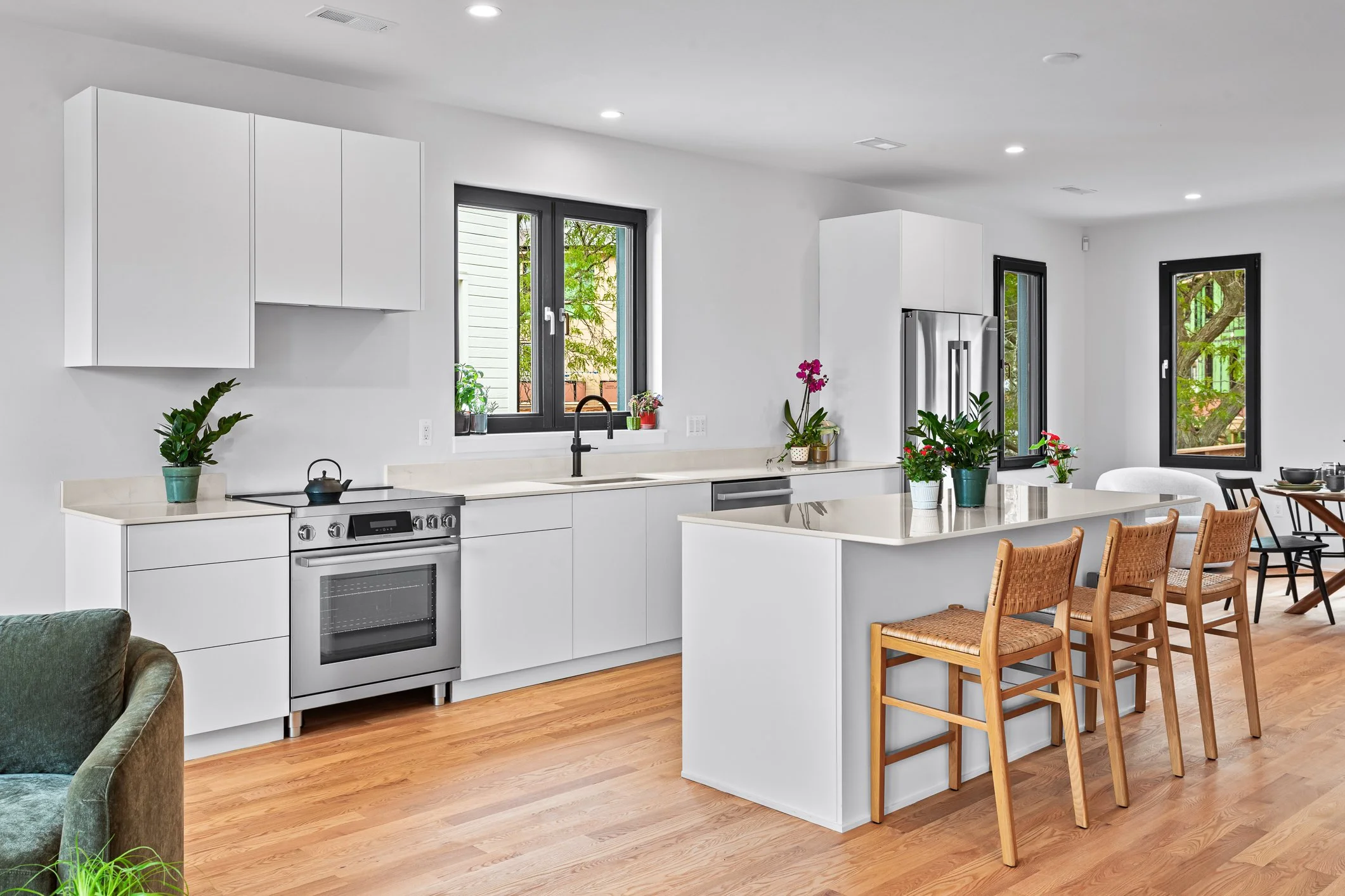 Modern kitchen with white cabinets, black window frames, stainless steel appliances, and wooden flooring. There is a kitchen island with potted plants, a dining area with black and wooden chairs, and trees visible through the windows.