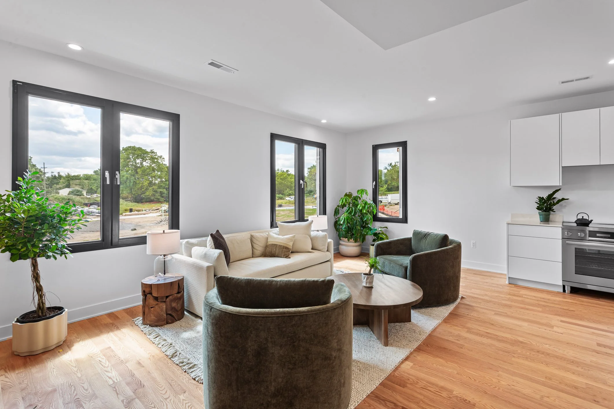 Modern living room with white walls, large windows, wooden flooring, a beige sofa, green and brown armchairs, small round wooden coffee table, potted plants, and a corner kitchen with white cabinets and stainless steel appliances.