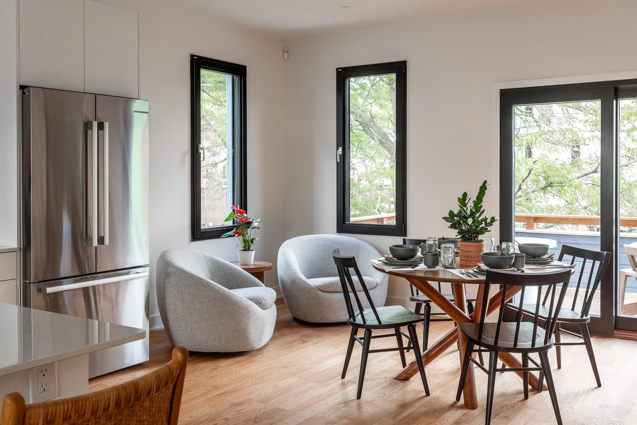 A modern dining area with a round wooden table set with black bowls, glasses, and utensils, surrounded by four black chairs. There are two white armchairs, a small wooden side table with a potted plant, and a large window and sliding glass door with 
