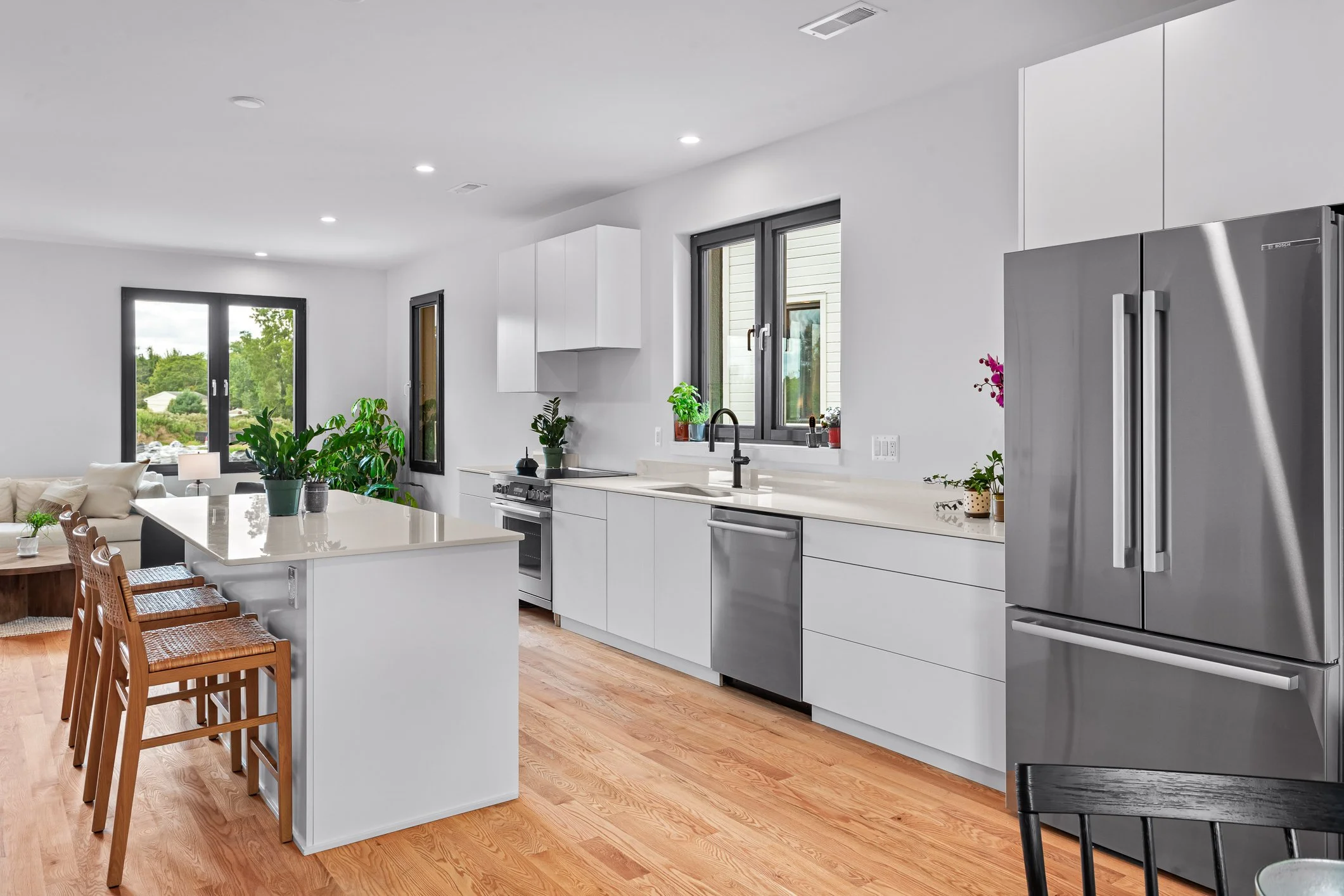 Modern kitchen with white cabinets, a large island, and hardwood floors. Stainless steel refrigerator, sink, and appliances. Windows with a view of greenery. Potted plants and minimal decor.