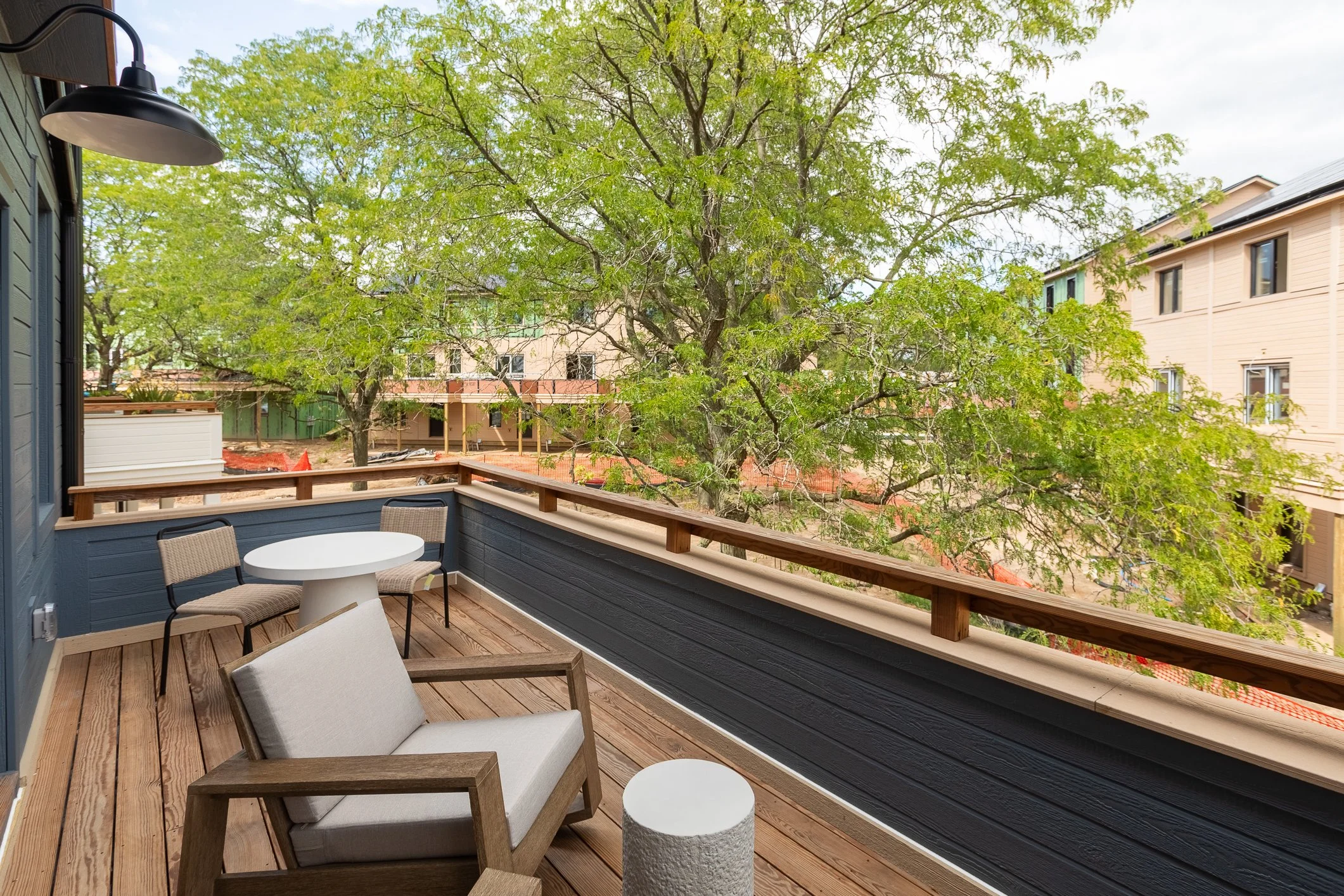View from a balcony with wooden flooring, an outdoor armchair, a small round table, and two chairs, overlooking a green tree and neighboring buildings.