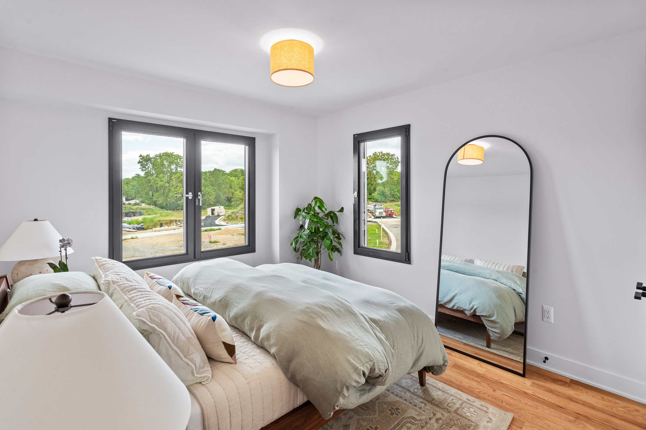 A bright bedroom with white walls, a large bed with light-colored bedding and pillows, a white bedside table with a white lamp, a tall mirror, a green potted plant, and two windows showing a view of greenery outside.
