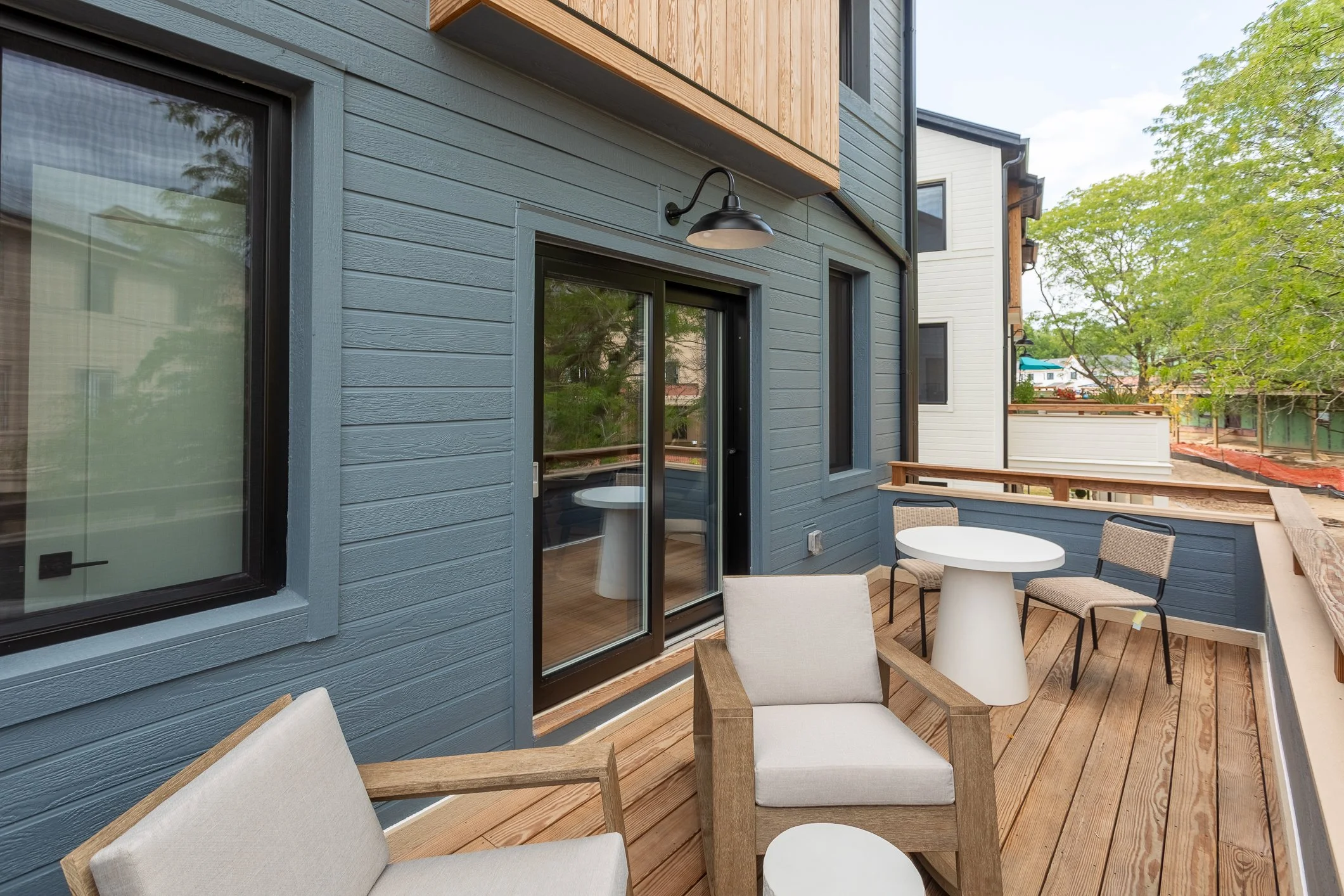 A modern outdoor balcony with wooden flooring, furnished with beige cushioned chairs, a white round table, and black chairs. The balcony is attached to a blue house with sliding glass doors and windows. There are trees and neighboring houses visible 