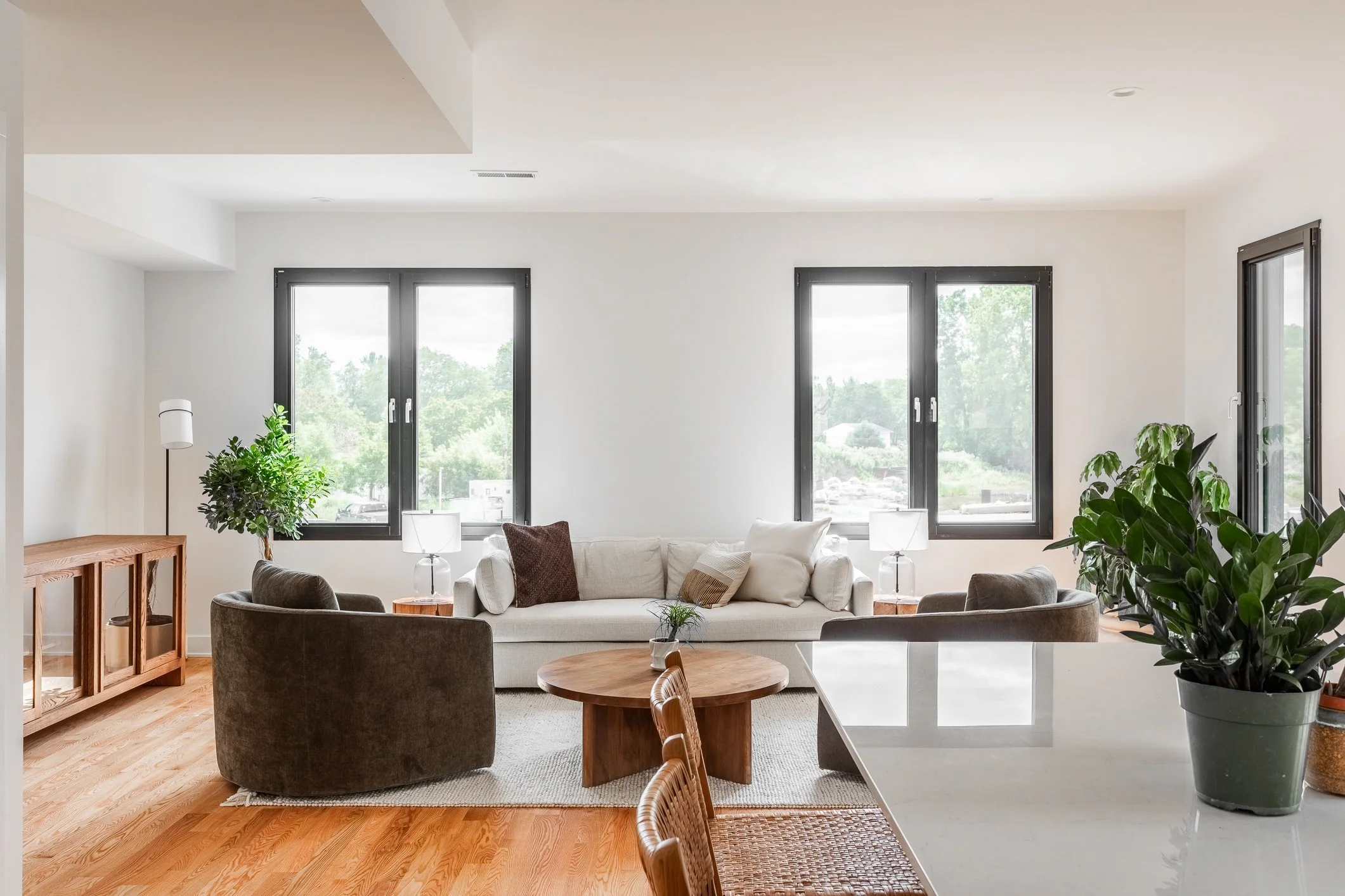 Living room with white walls, three large windows, a white sofa with pillows, two brown armchairs, a wooden coffee table with a small potted plant, side tables with lamps, green plants, and a wooden sideboard.