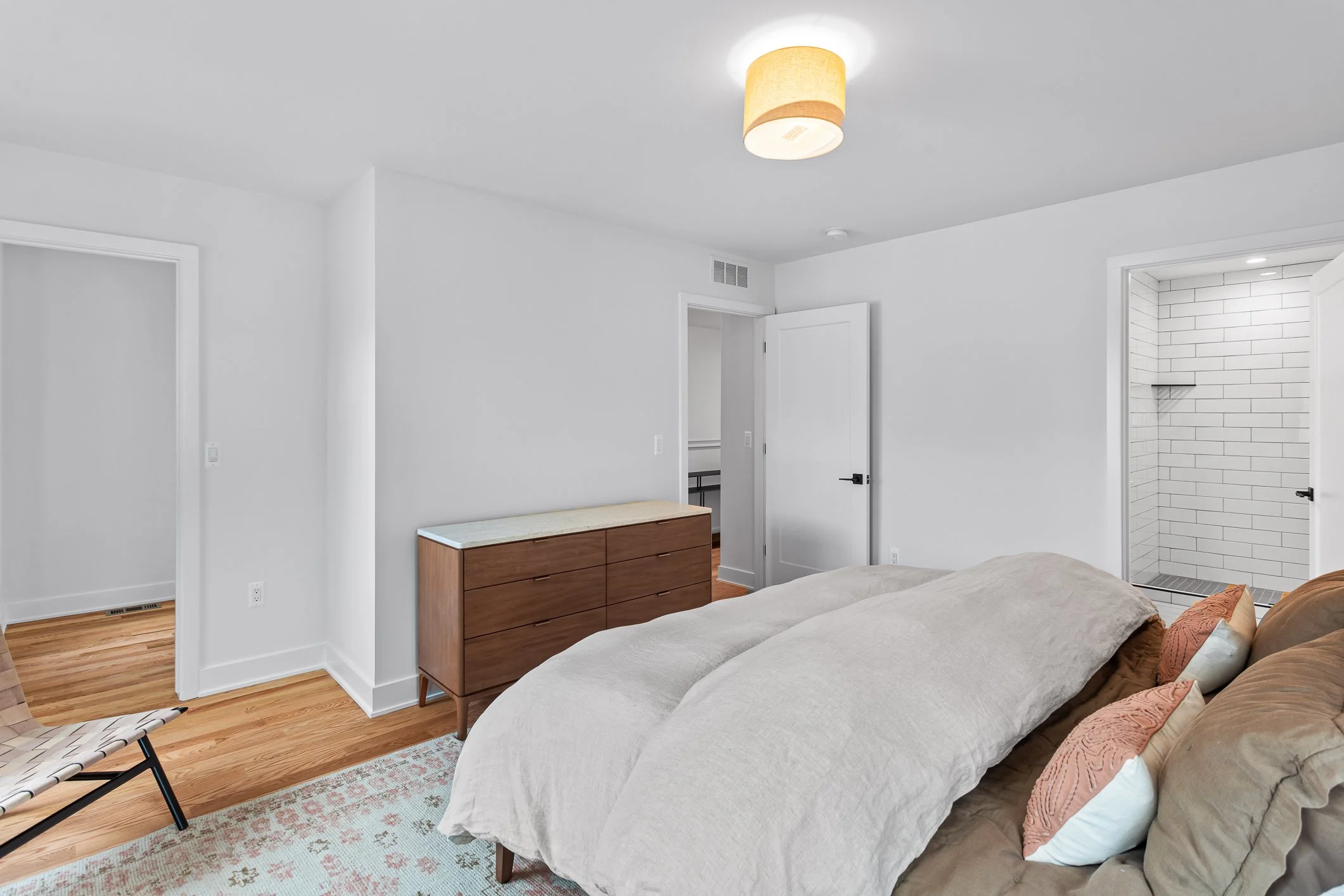 A bedroom with white walls, a bed with beige bedding and pillows, a wooden dresser, and a doorway leading to a tiled bathroom with a walk-in shower.