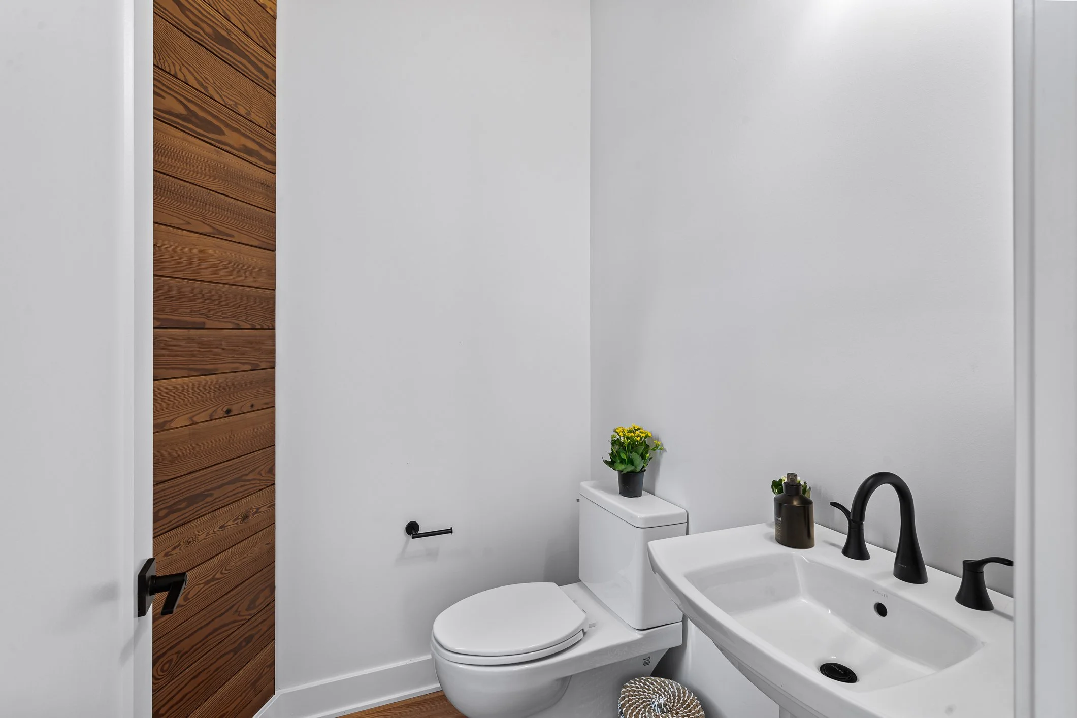 A minimalist bathroom with white walls, a white toilet with a black plant on top, a black toilet paper holder, and a white sink with black fixtures. There is a wooden door on the left and a small basket on the floor.