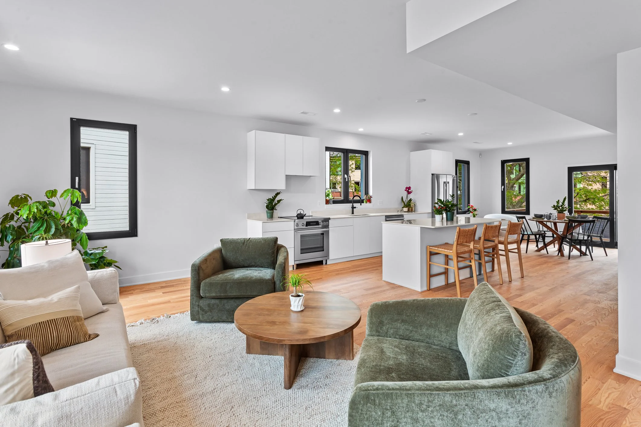 Open-concept living room and kitchen with white walls, wooden floors, large windows, green and beige furniture, and indoor plants.