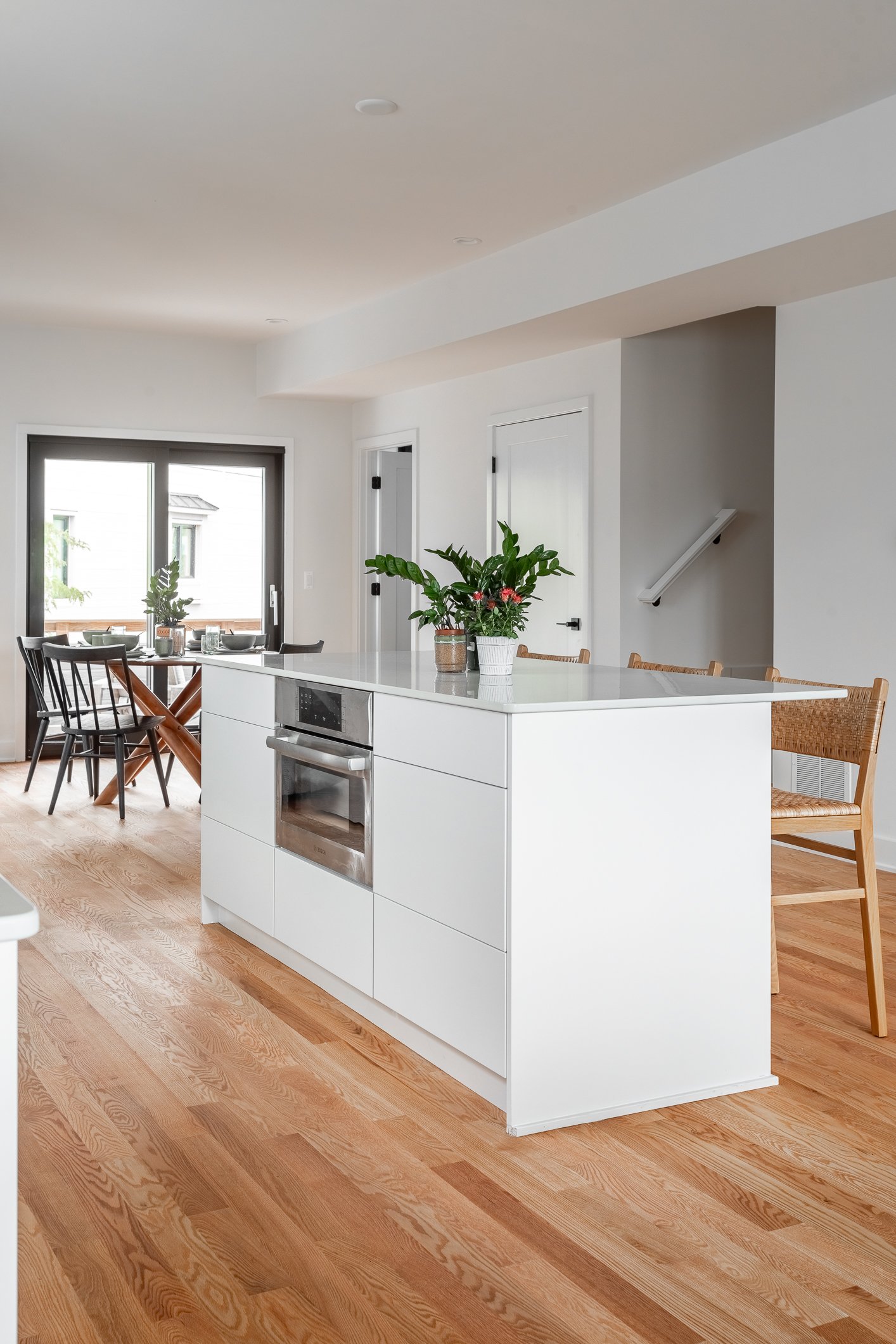 Modern kitchen with white island, wooden floors, potted plants on the counter, dining area with black chairs, and sliding glass door leading outside.