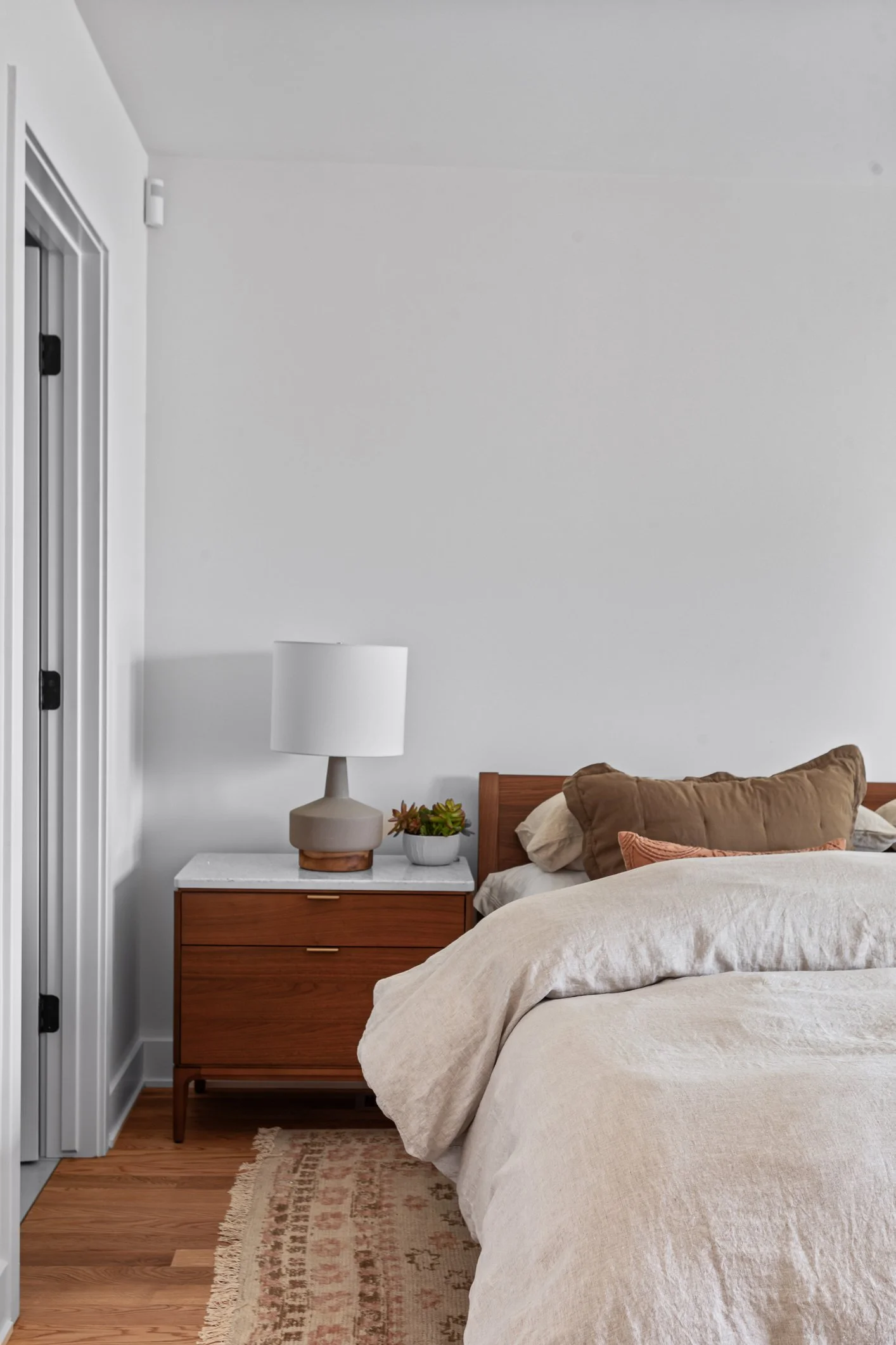 A bedroom scene with a wooden nightstand, a white table lamp, a small potted plant, a bed with beige and brown pillows, and a white wall background.