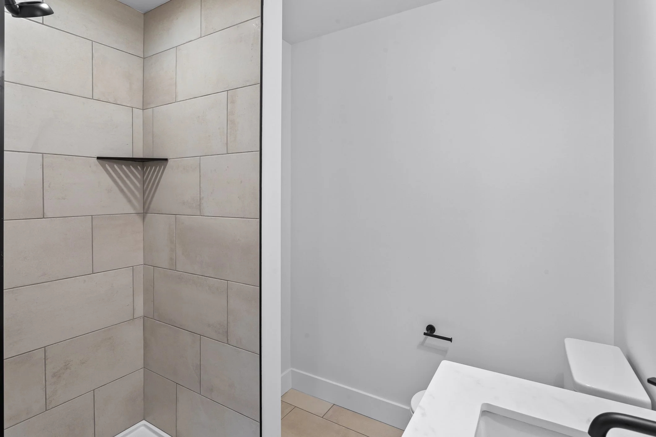 Empty bathroom with a walk-in shower with beige tiles and a black showerhead, a white toilet with a black toilet paper holder, and a white marble vanity with a black faucet.