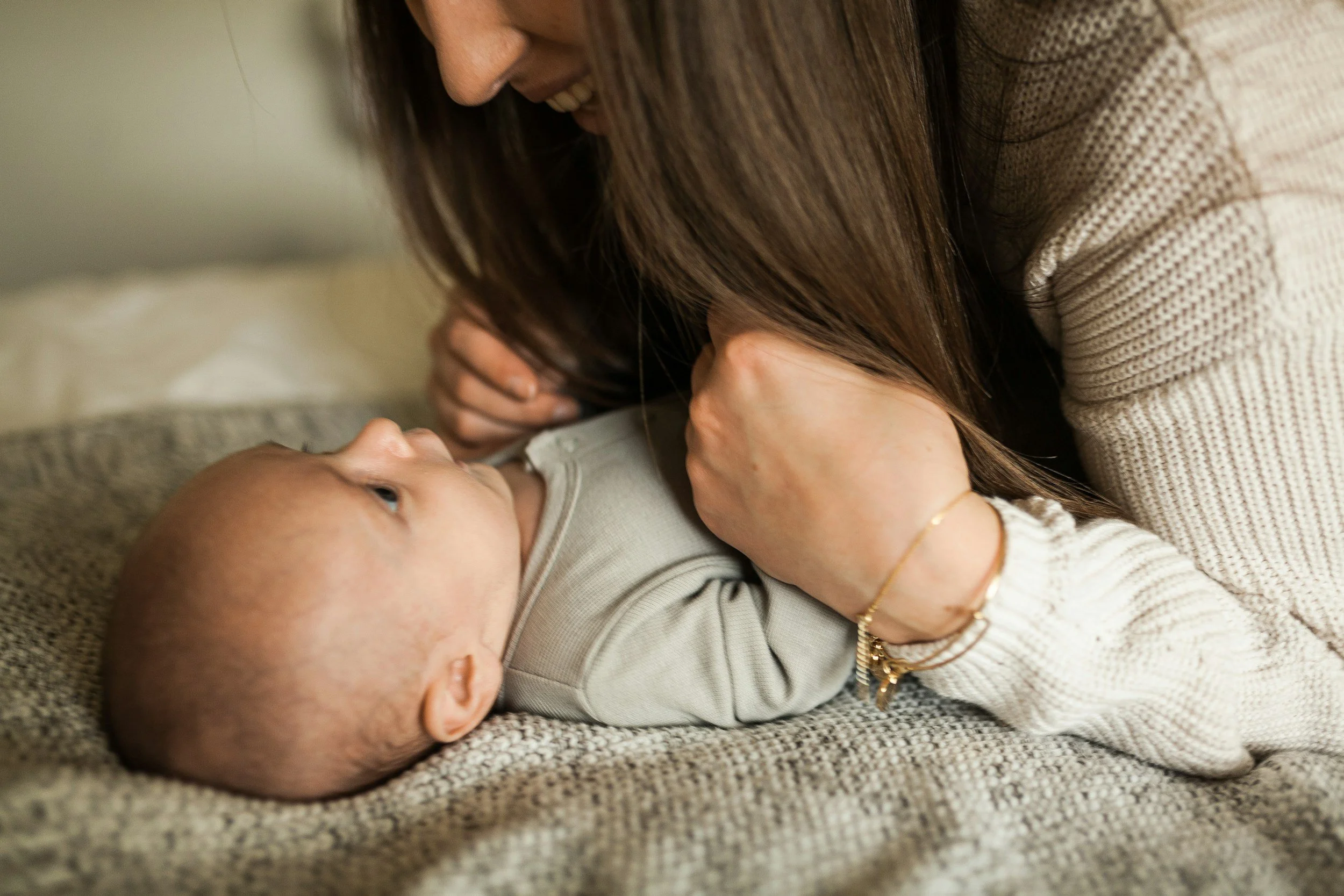 A woman and baby lying on a textured brown carpet, close to each other, smiling, and touching. The woman has long dark hair and is wearing a beige sweater. The baby has light skin, a round head with few hair, and is wearing a light-colored outfit.