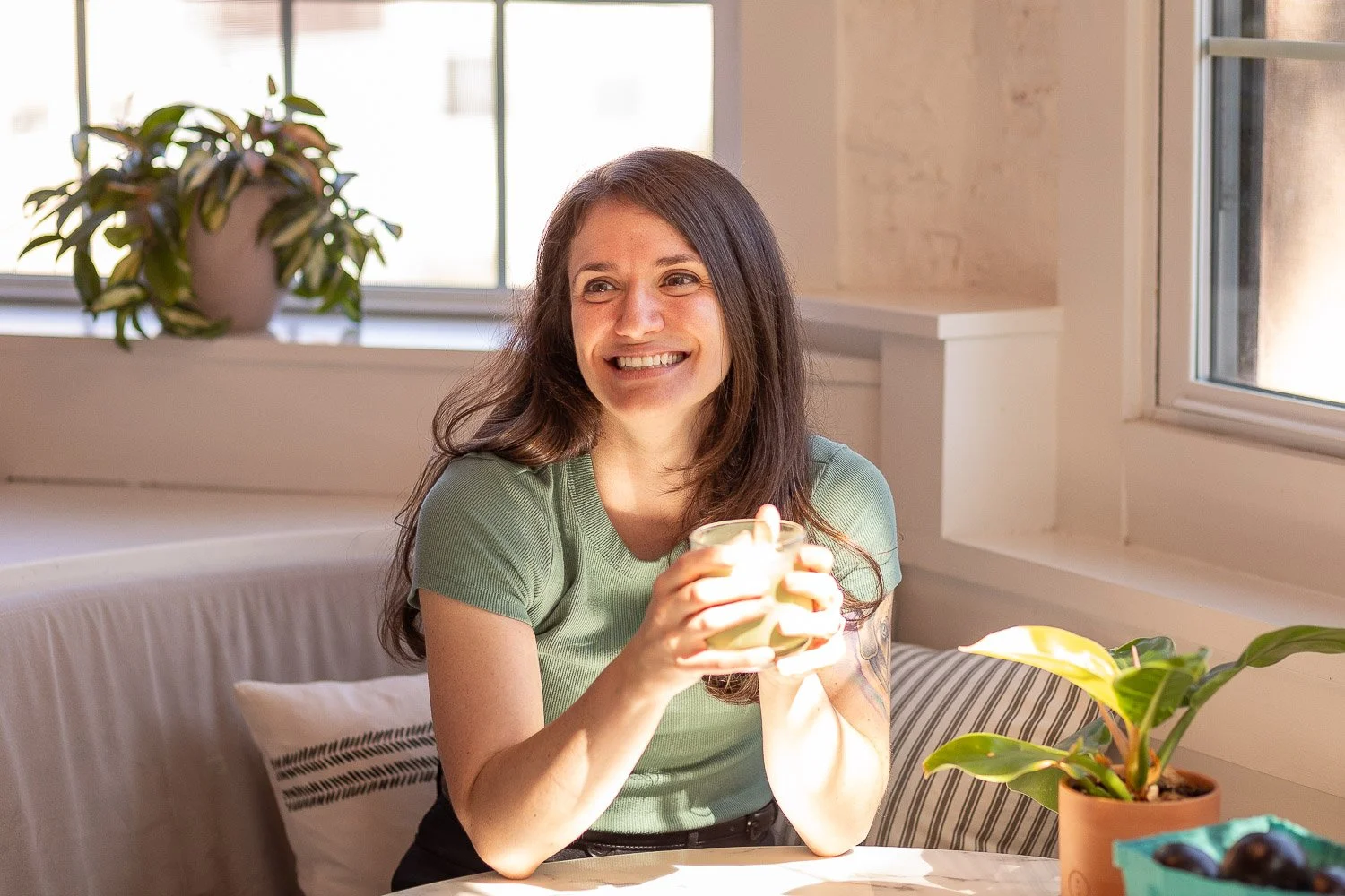 A woman with long dark hair smiling and holding a cup seated at a table near windows with potted plants on the window sills.