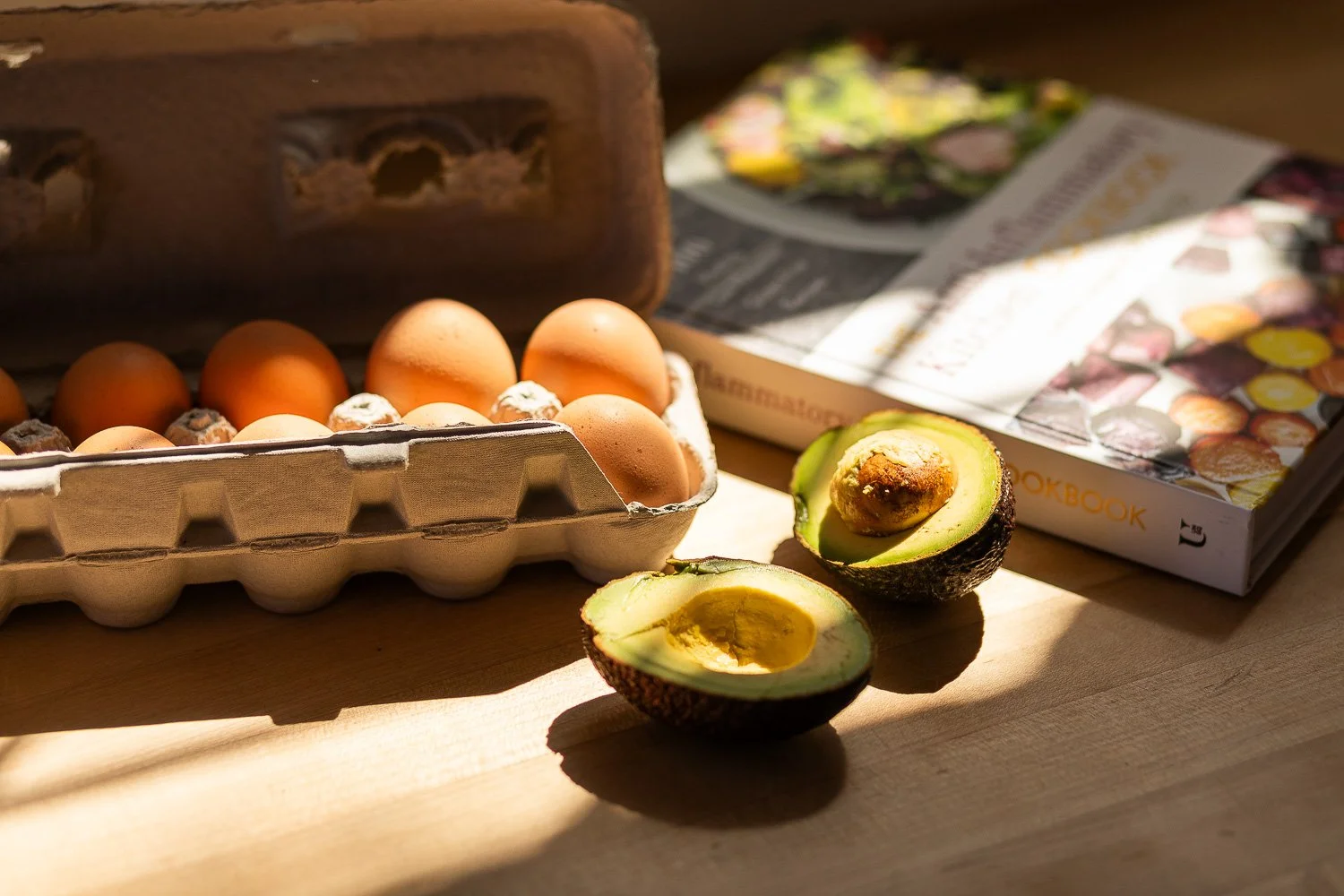 Egg cartons with brown eggs, halved avocado with seed, and a gardening book on a wooden surface illuminated by sunlight.