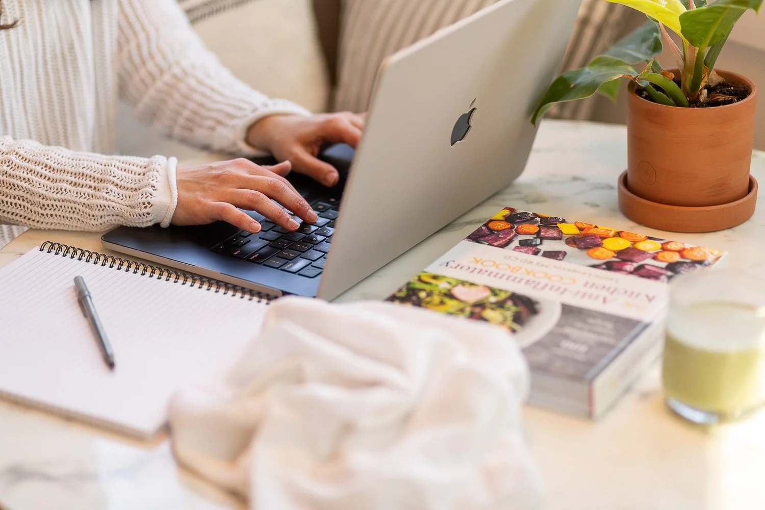 Person typing on a silver MacBook laptop on a marble table, with a notepad and pen nearby, a potted plant, some colorful magazines, and a glass of water.