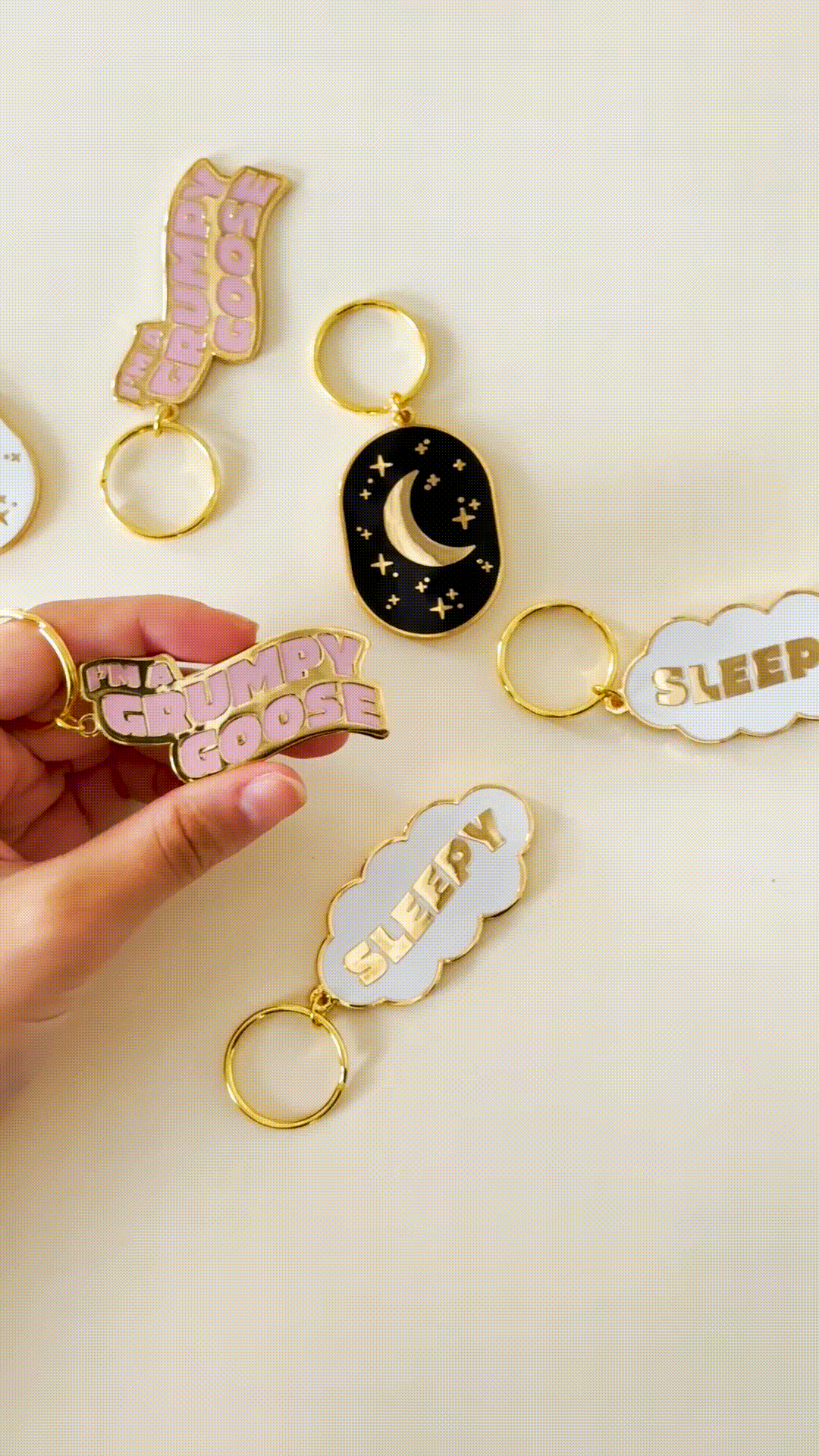 A hand holds three unique keychains against a tan background: a white "Sleepy Cloud," "Grumpy Goose" with pink wavy text, and an oval "Night Sky Moon and Stars" design.