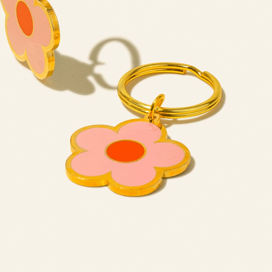 Close-up of a pink flower keychain with a red center, highlighting its gold metal detailing, lying on a light background.