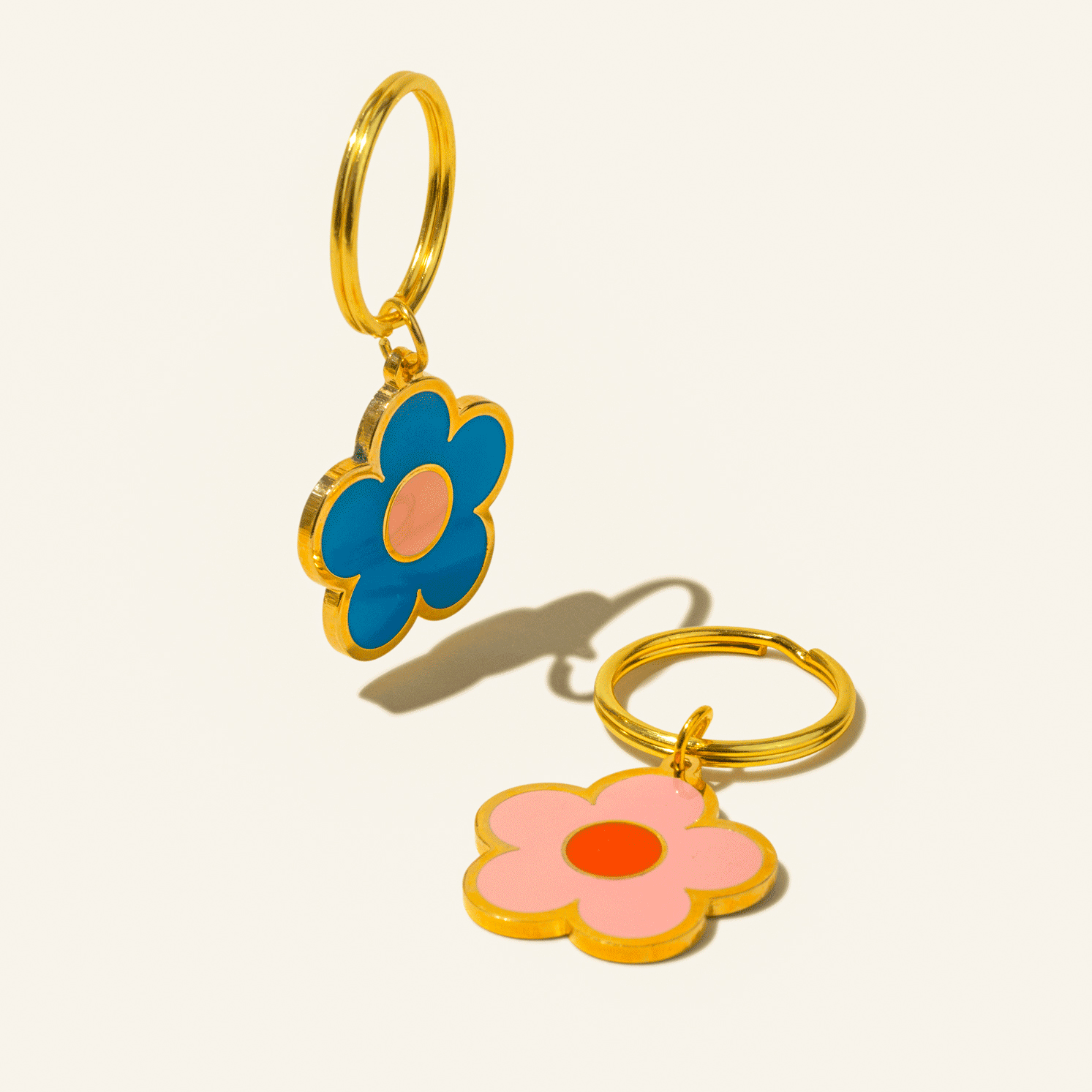 Two flower-shaped keychains with gold rings, one in blue with a pink center and the other in pink with an orange center, displayed on a light background with soft shadows.