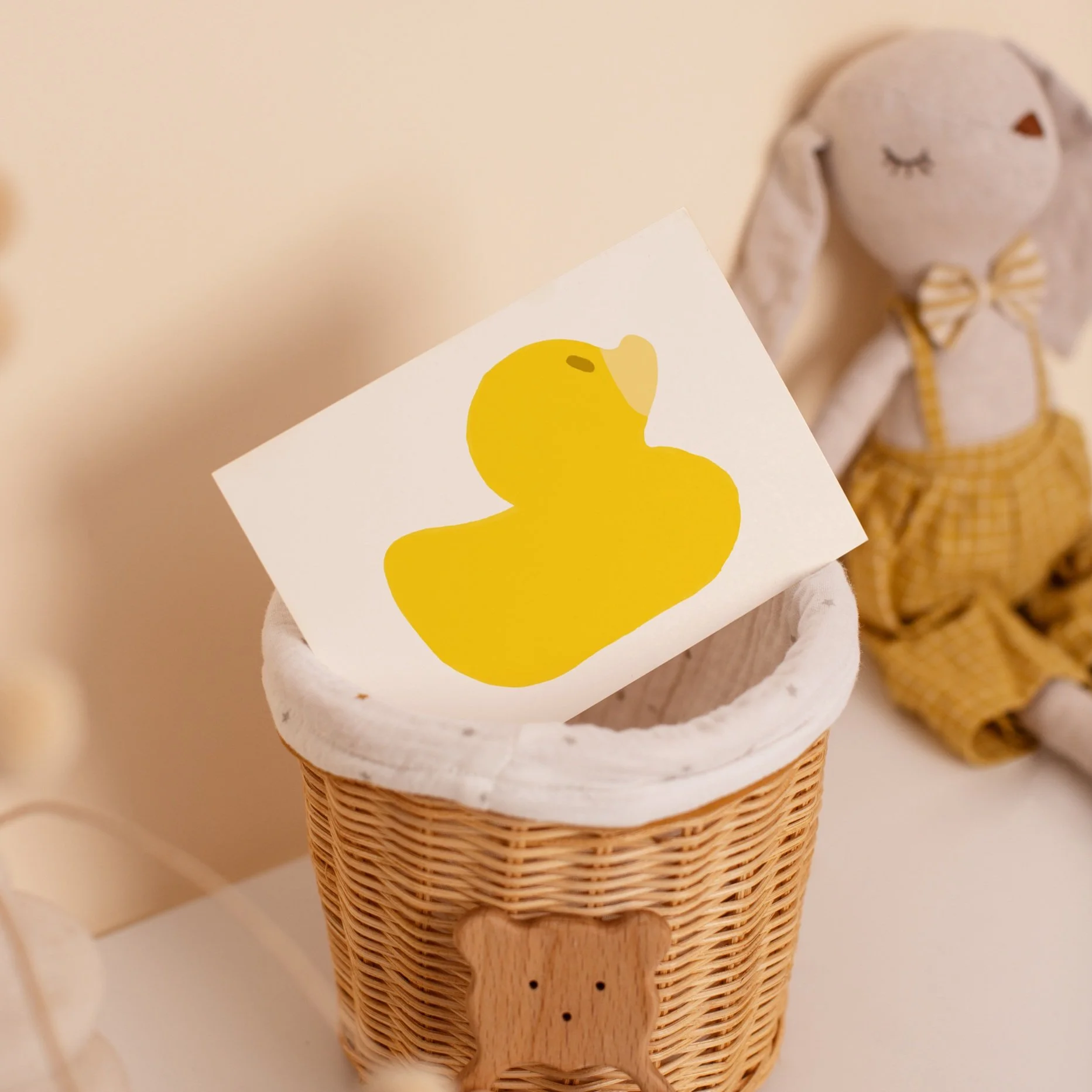 Greeting card featuring a bold yellow rubber duck design, placed in a wicker basket among soft, neutral-toned baby room decor.
