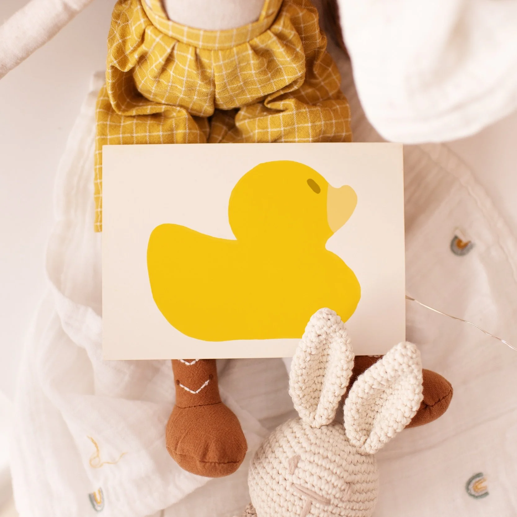 Greeting card with a large yellow rubber duck illustration, displayed alongside baby-themed decorations including plush toys and a soft muslin blanket.
