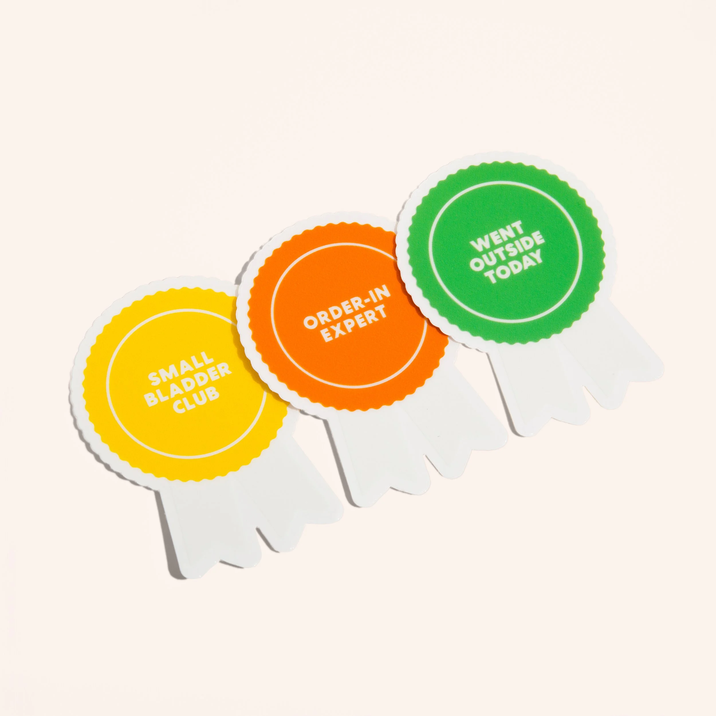 Set of three award-shaped stickers in bright colors with humorous text: "Small Bladder Club" in yellow, "Order-In Expert" in orange, and "Went Outside Today" in green.