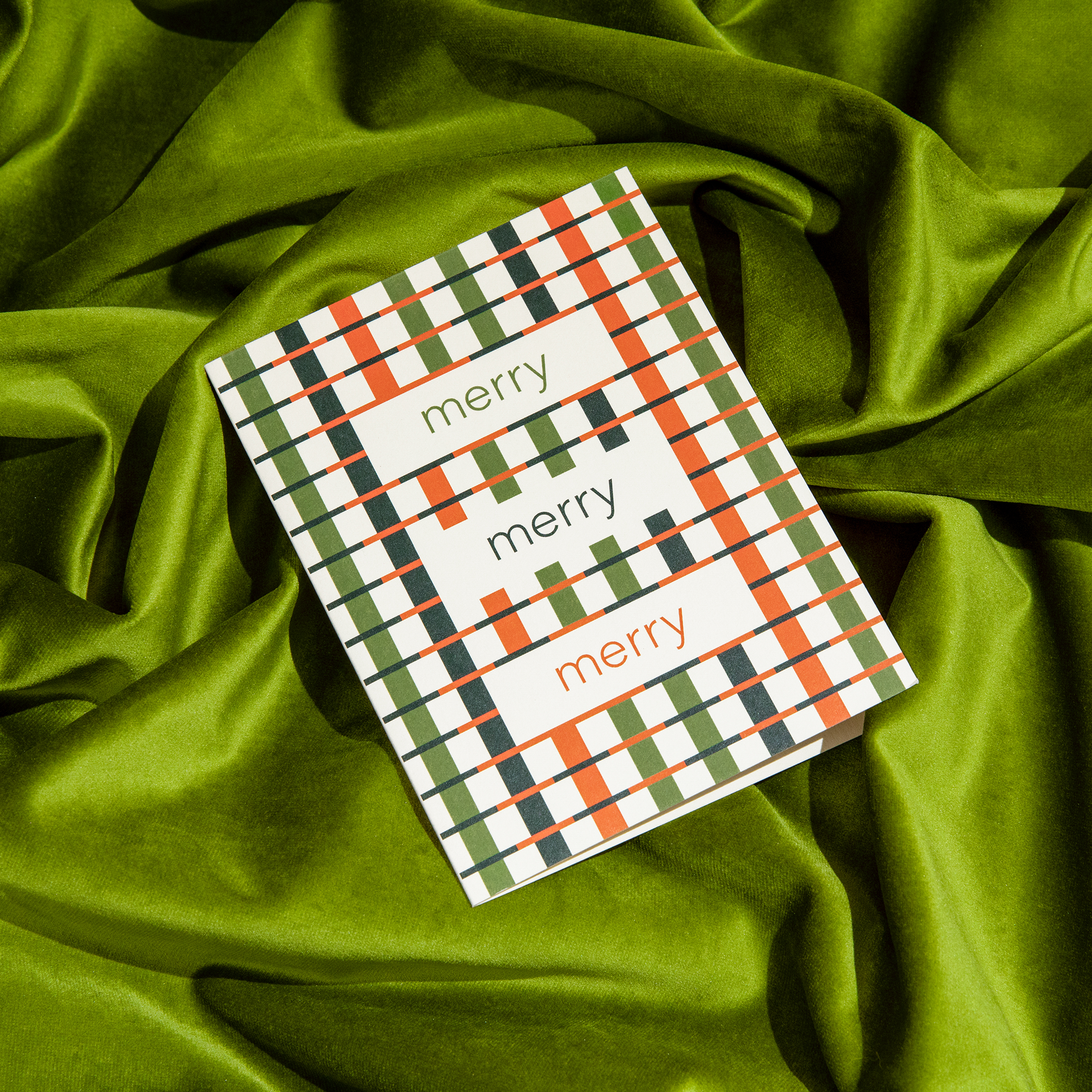 Holiday greeting card with a geometric red, green, and black pattern and the repeated text 'merry merry merry,' placed on a rich green velvet fabric backdrop.