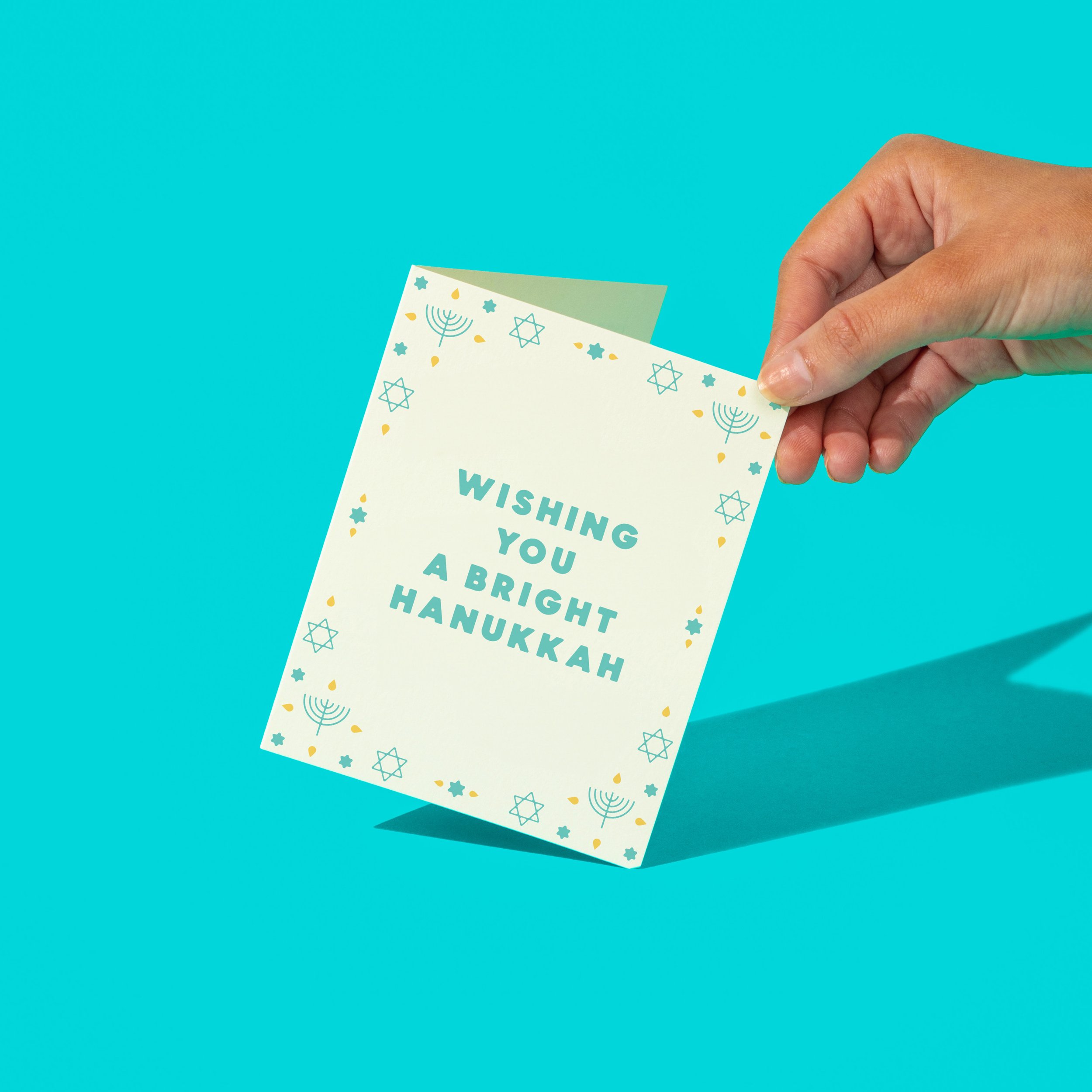 A hand holding a greeting card with the message 'Wishing You a Bright Hanukkah' in teal text, surrounded by menorah and Star of David symbols. The card is set against a vibrant blue background.