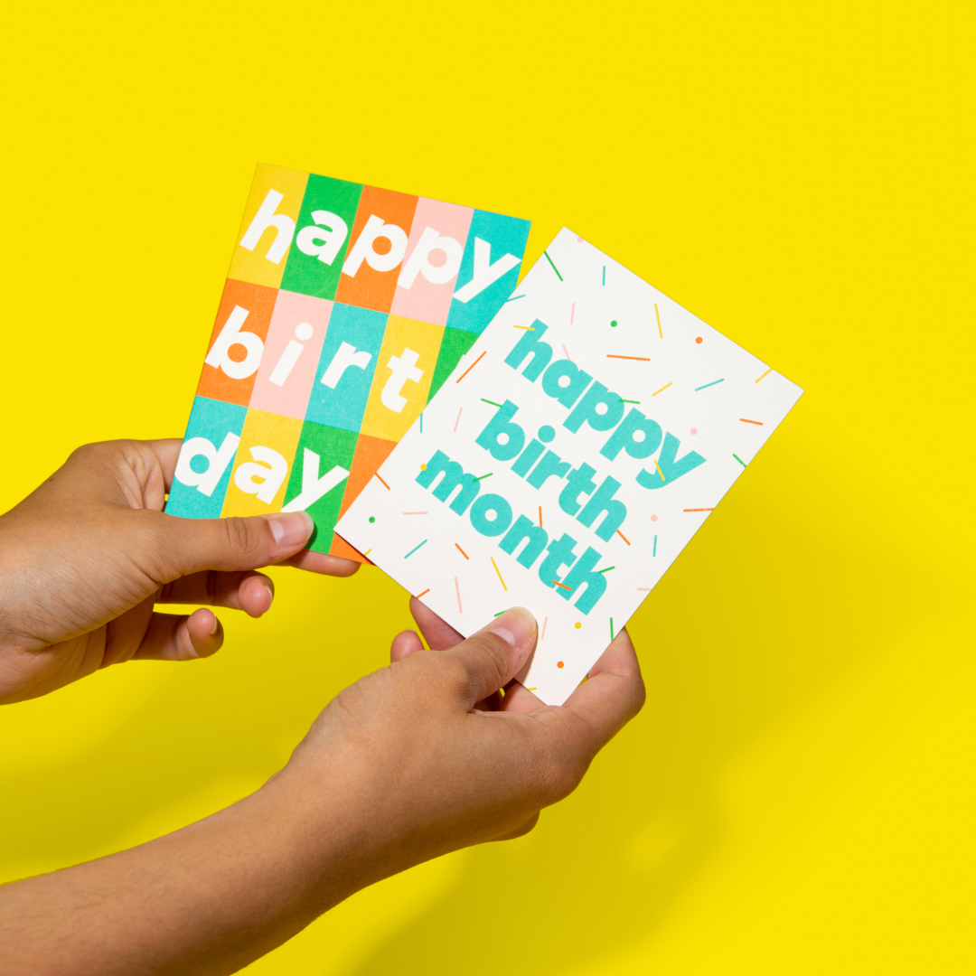 Hands holding two colorful birthday cards, one with the text "happy birthday" in a multicolored grid pattern, and the other with "happy birth month" and confetti accents, against a bright yellow background.