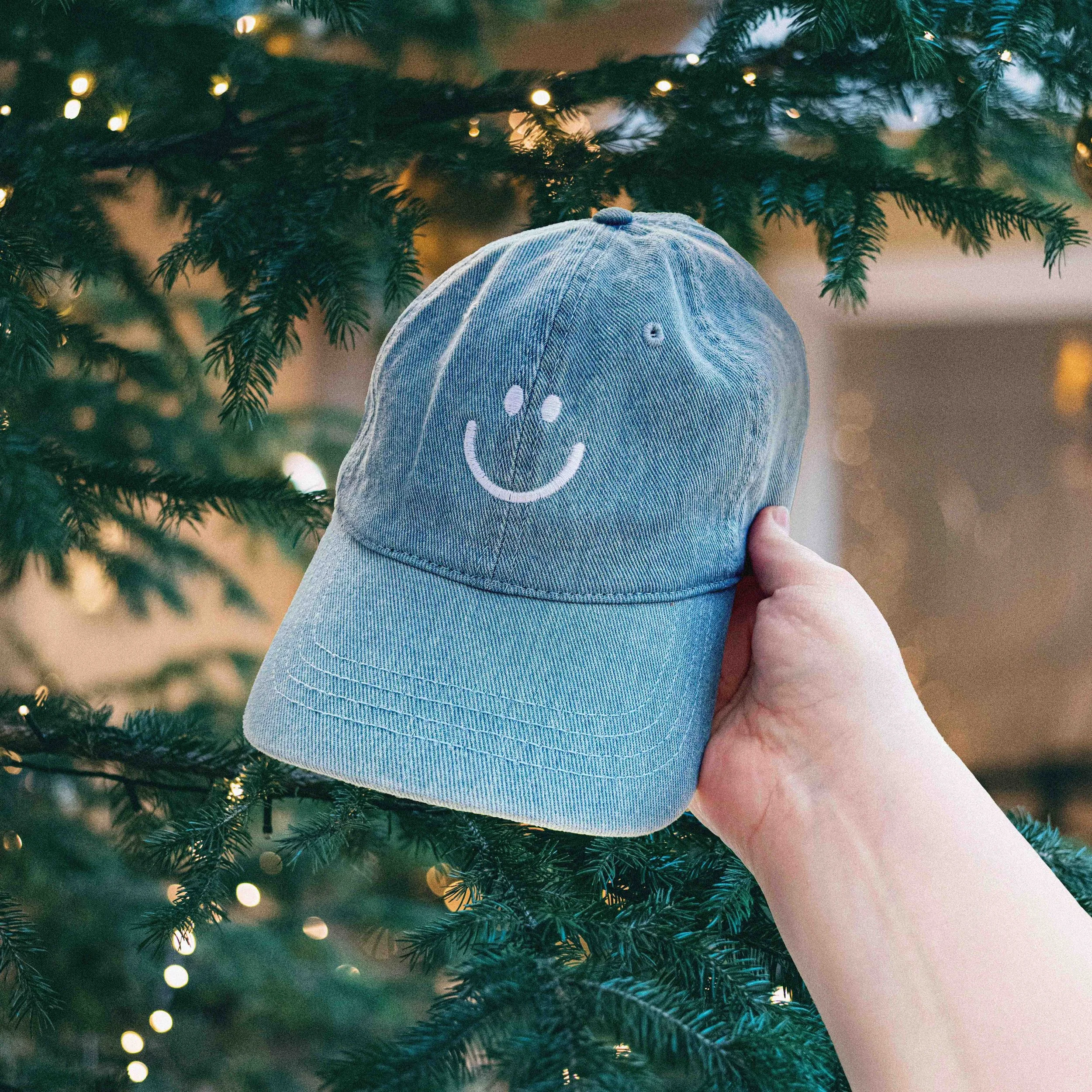 Hand holding a denim baseball cap with a simple embroidered happy face, in front of a Christmas tree adorned with string lights.