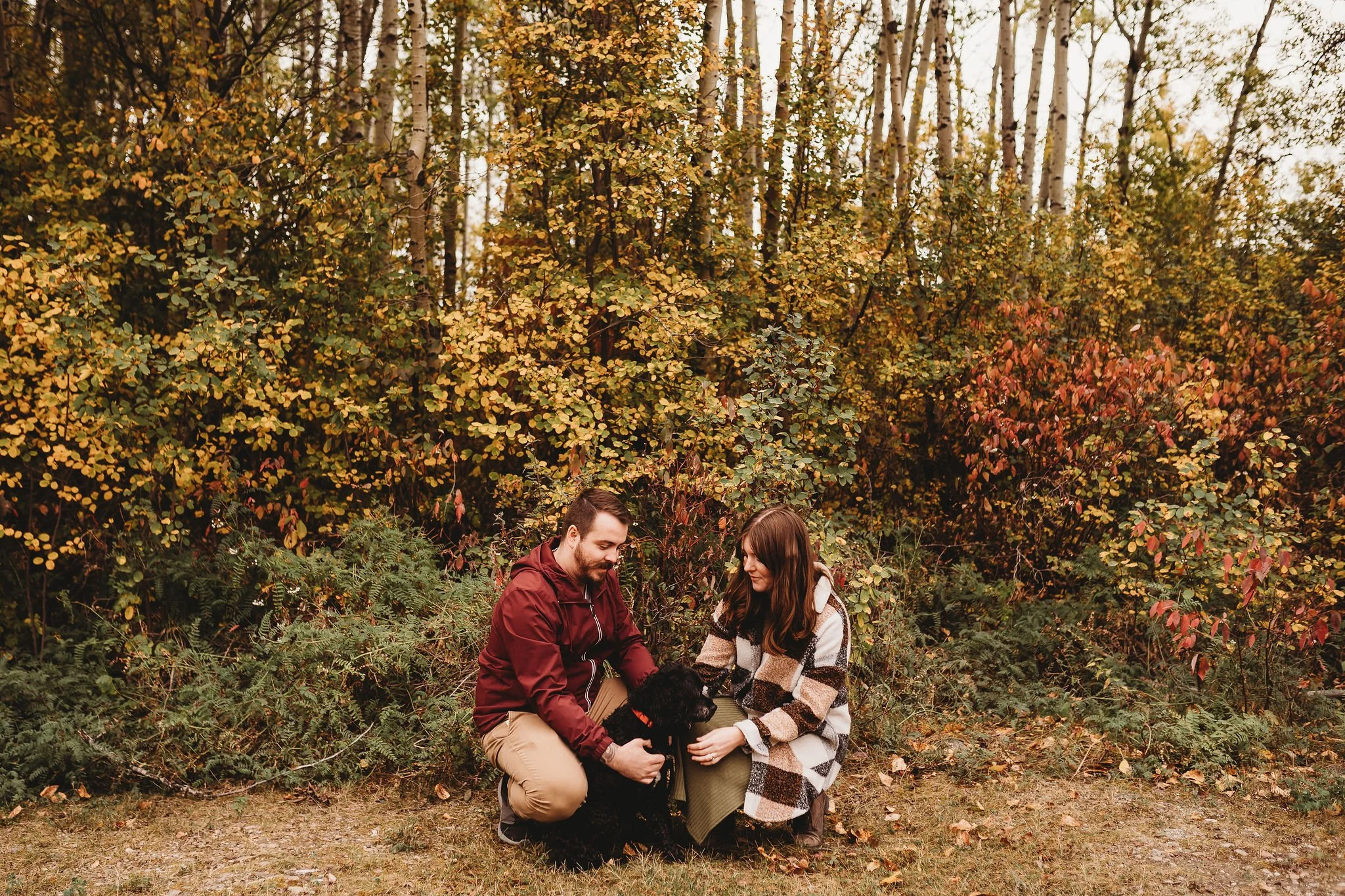 A man and woman kneeling outdoors amidst autumn foliage with a black dog.