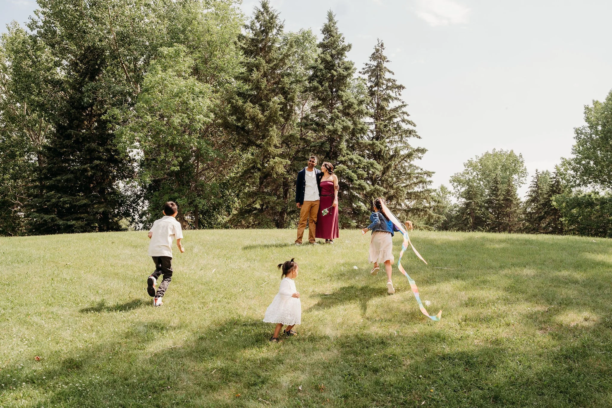 A family of five children playing on a grassy hill under the sun, with two adults standing together nearby and surrounded by tall trees.