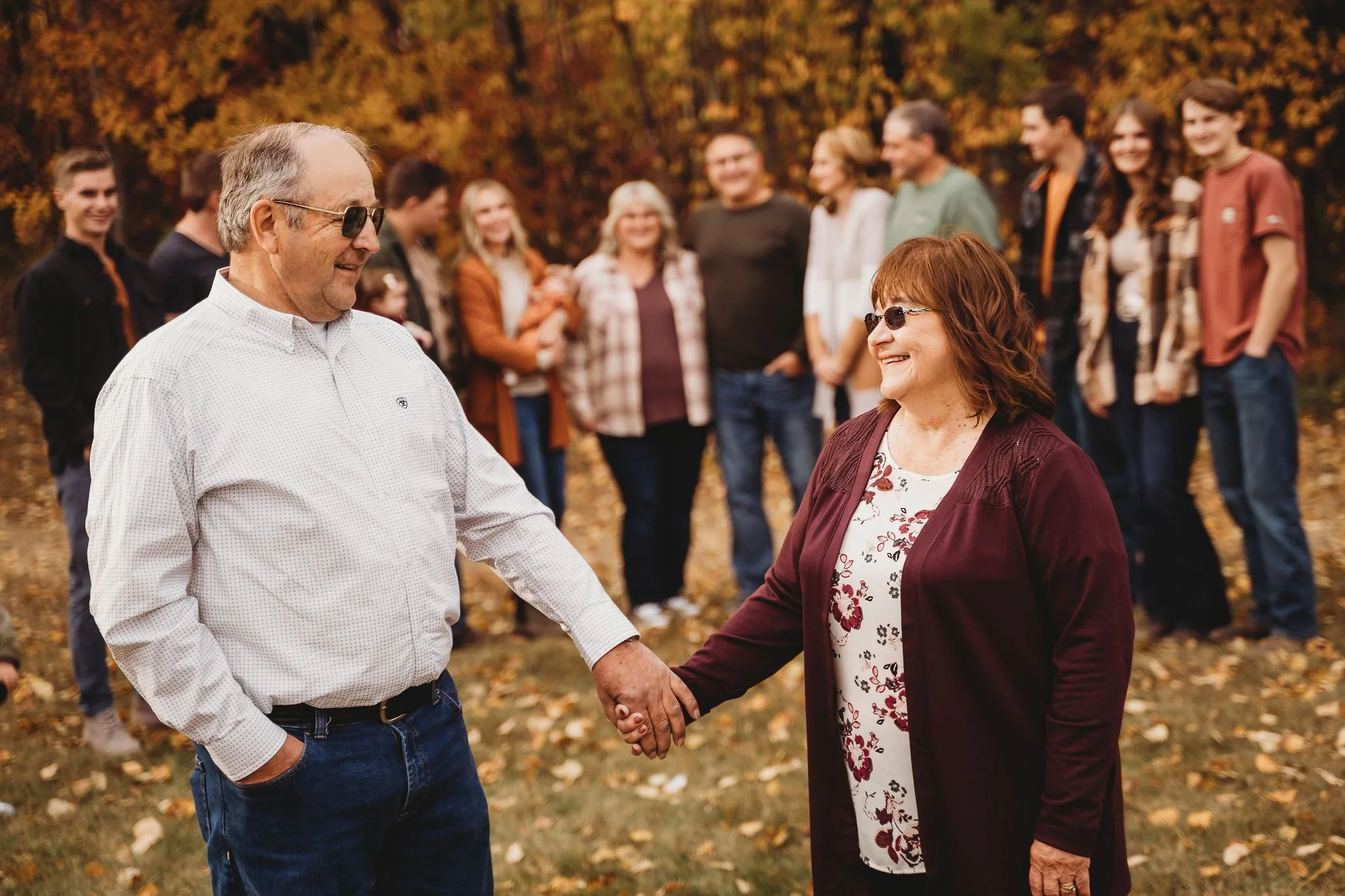 An elderly man and woman holding hands and smiling at each other outdoors with a group of people in the background during fall.