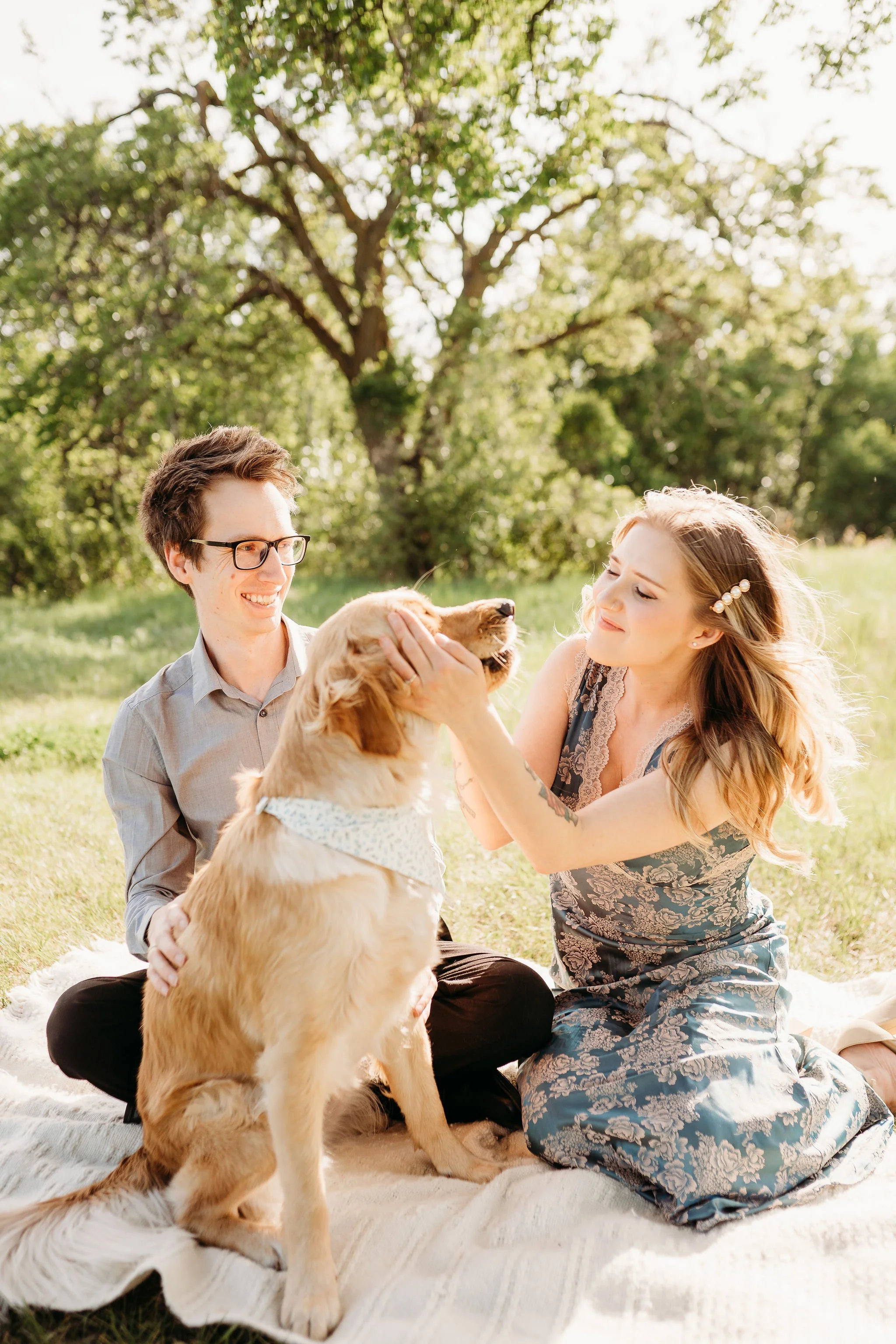 A couple and their dog sitting on a blanket outdoors under a large tree on a sunny day, the woman gently holding the dog's face.