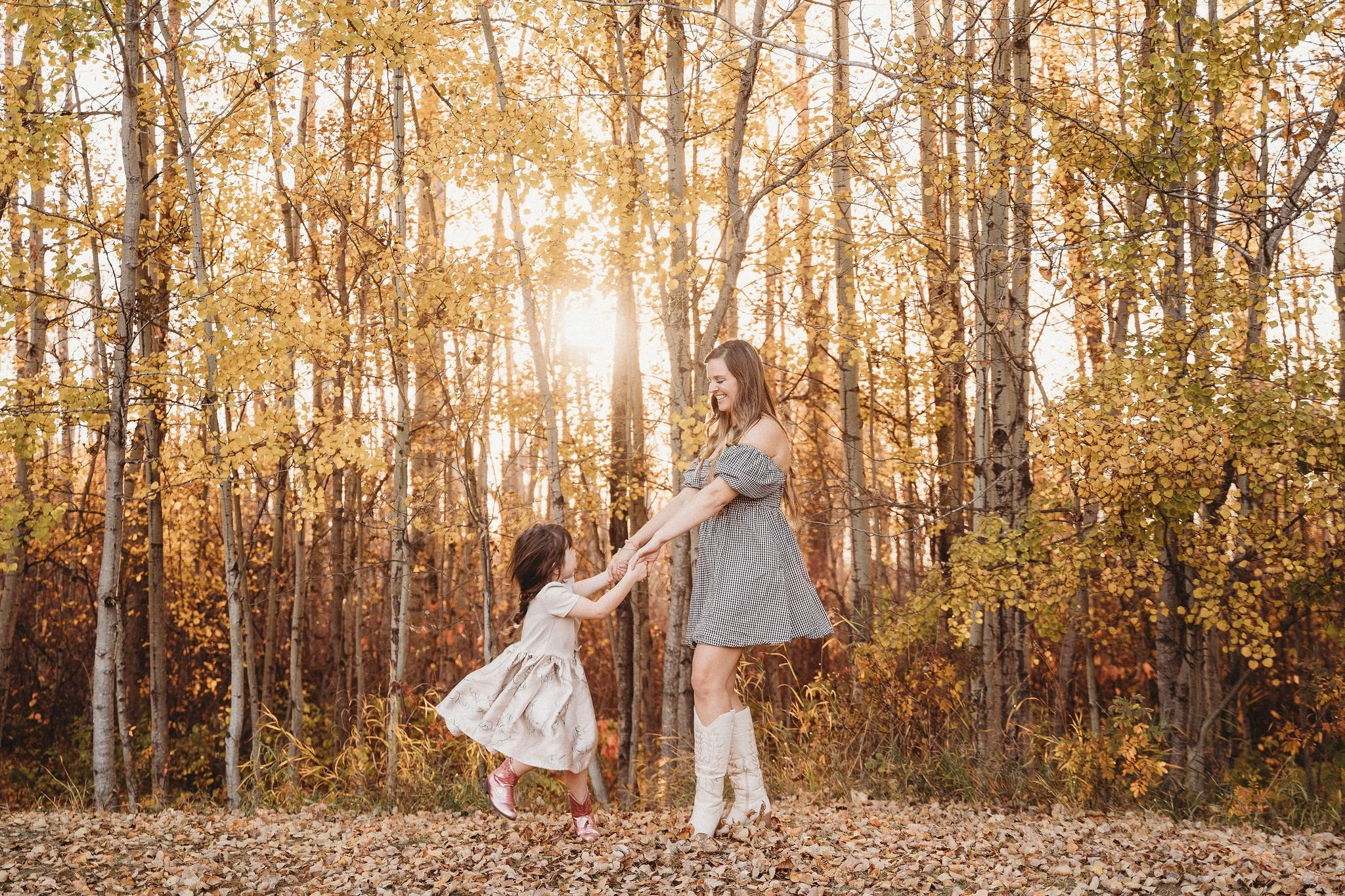 A woman and a young girl are playing and dancing in an autumn forest. The woman appears to be spinning the girl around, both are smiling. Sunlight filters through the golden leaves of the trees.