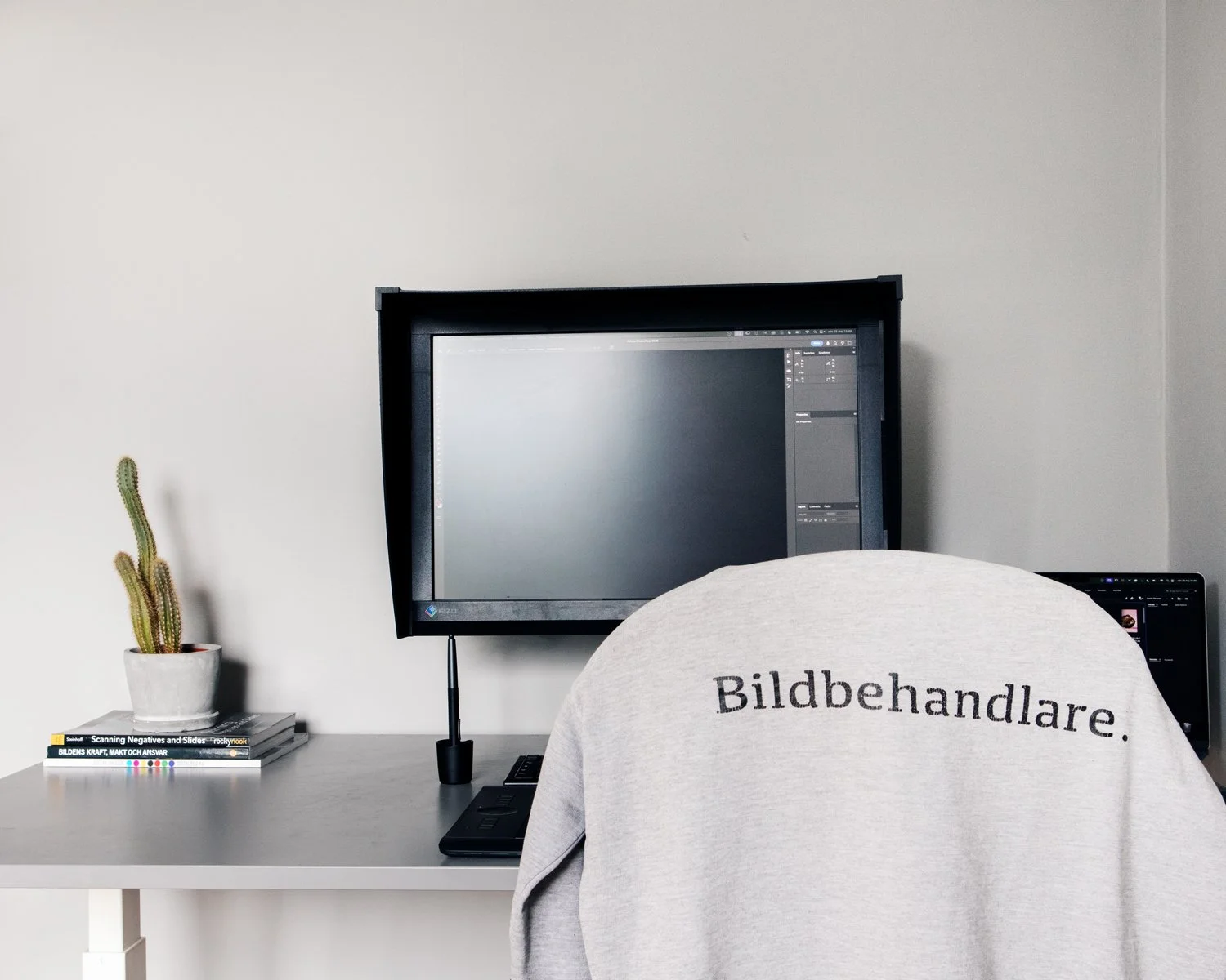 A desk setup with a cactus in a white pot, a stack of books, a computer monitor displaying editing software, a graphics tablet, and a person wearing a gray sweatshirt with 'Bildbehandlare' written on the back, facing the computer.