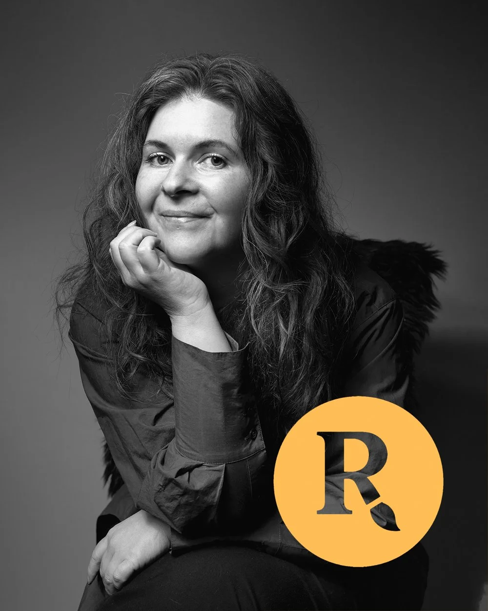 Black-and-white portrait of a woman with long curly hair, resting her chin on her hand, wearing a dark jacket with a fur collar, with a yellow circle containing a black letter 'R' and a leaf icon in the lower right corner.