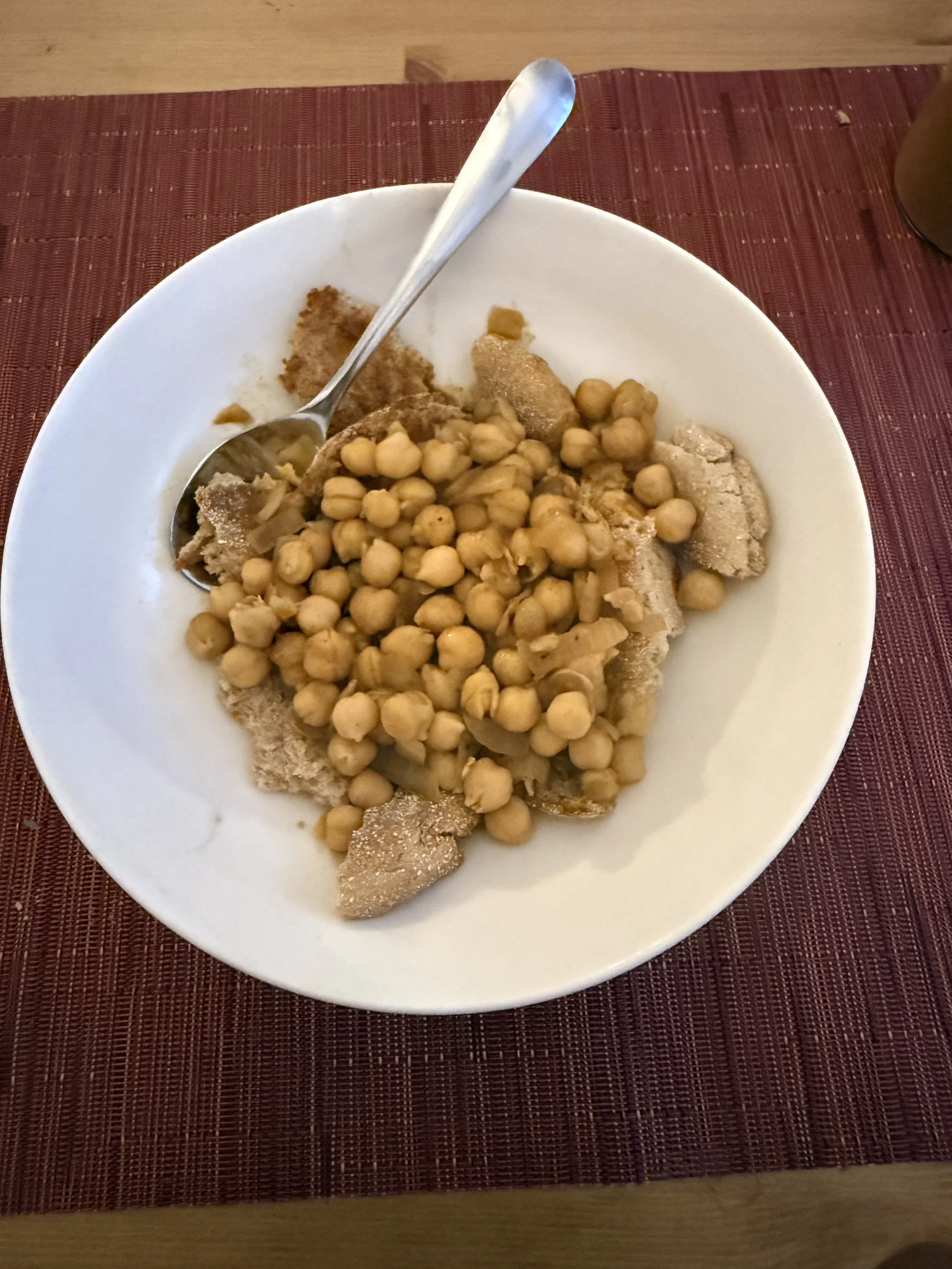 A white bowl filled with chickpeas and sliced tripe, with a spoon resting inside, placed on a red placemat on a wooden table.