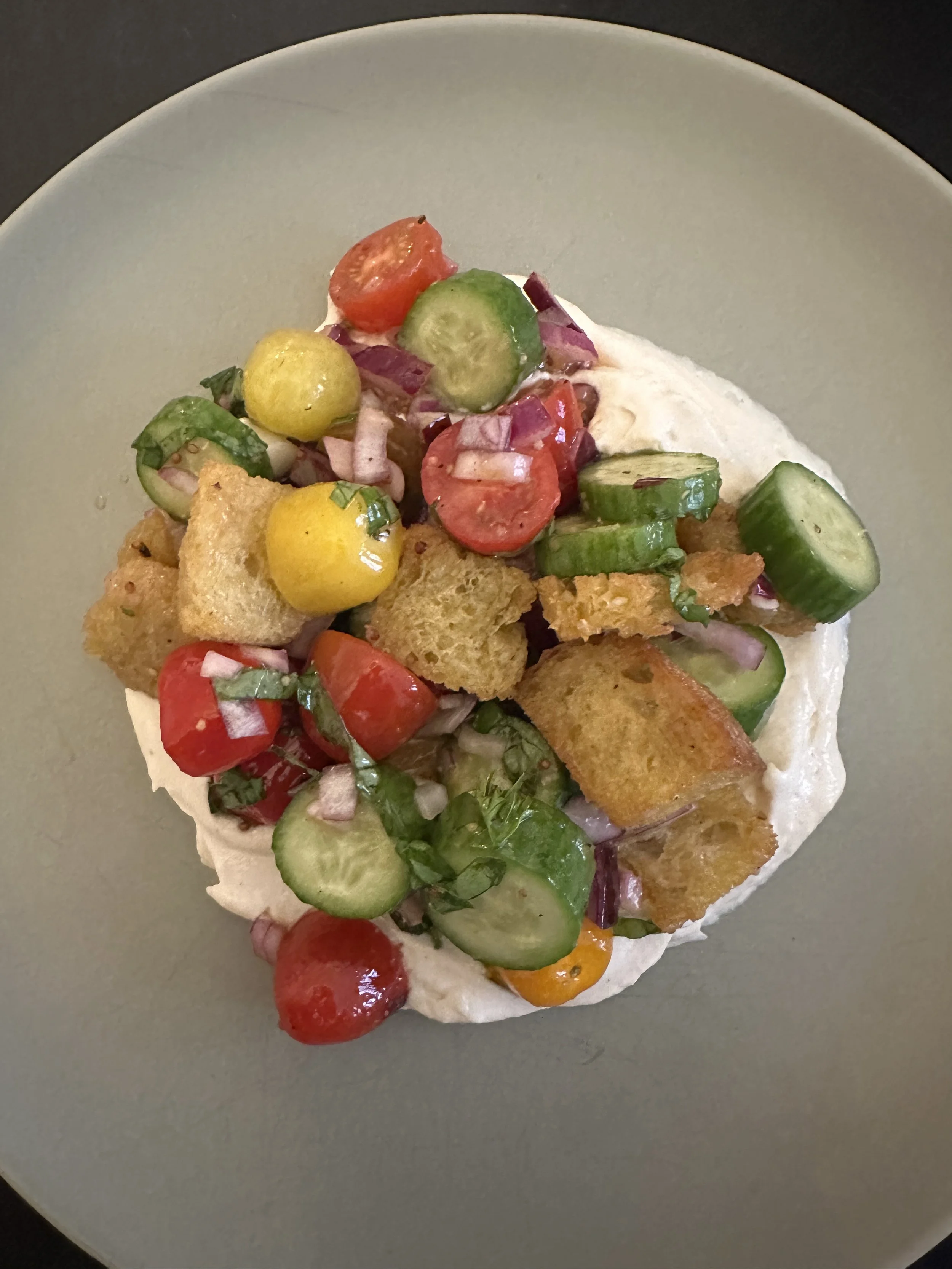 A close-up of a bowl of salad topped with cherry tomatoes, cucumbers, red onions, croutons, and chopped herbs on a gray plate.