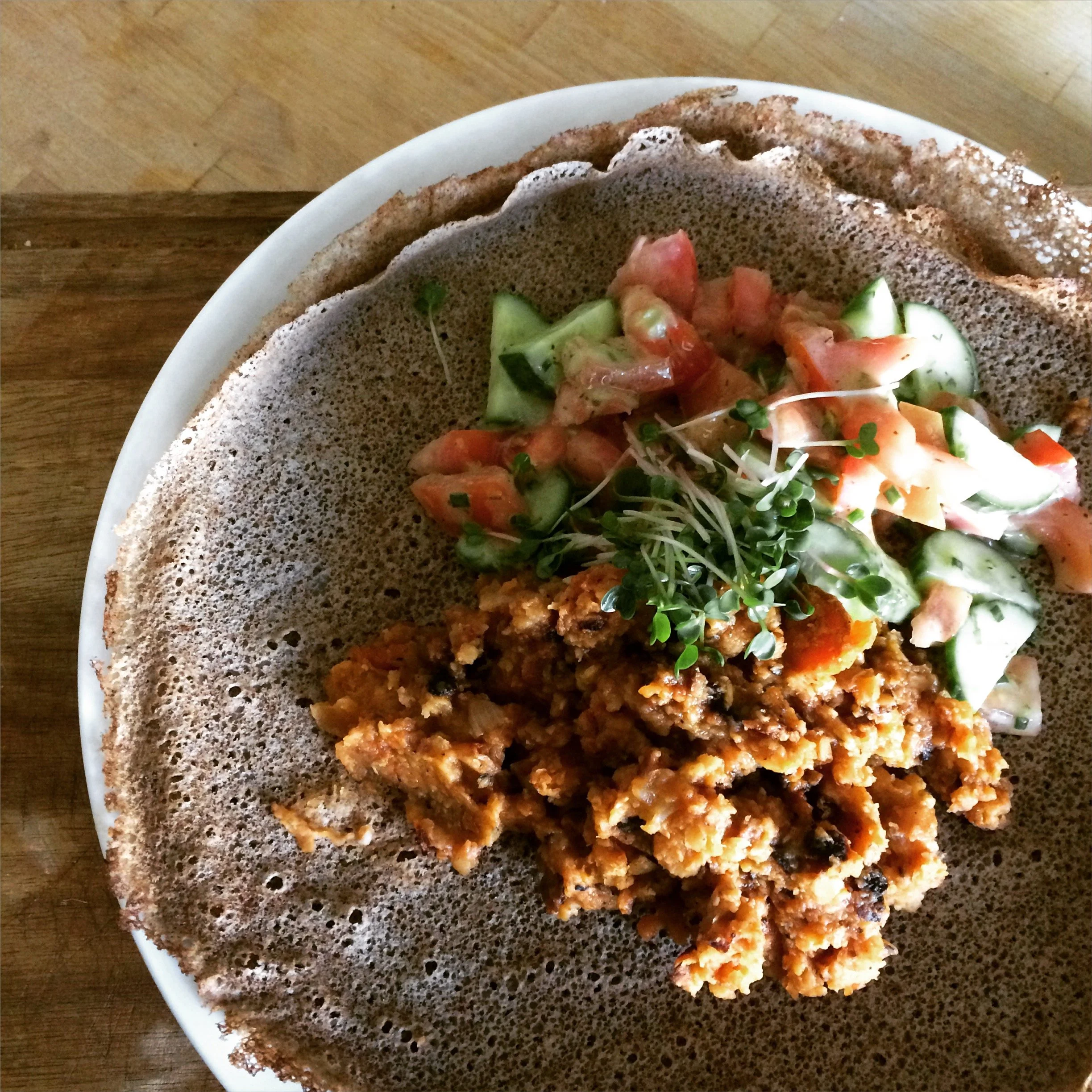 A plate with a large, thin, crispy injera bread topped with a vegetable salad, spicy lentil stew, and microgreens.