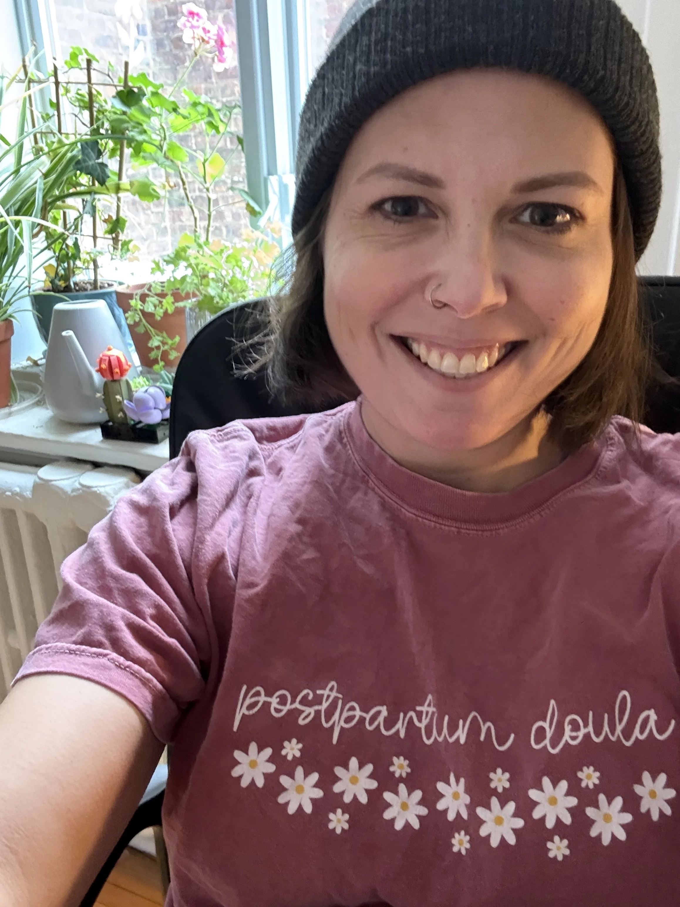 A smiling woman with short brown hair wearing a black beanie and pink t-shirt with white text and daisy designs, taking a selfie indoors near a window with potted plants and flowering plants in the background.