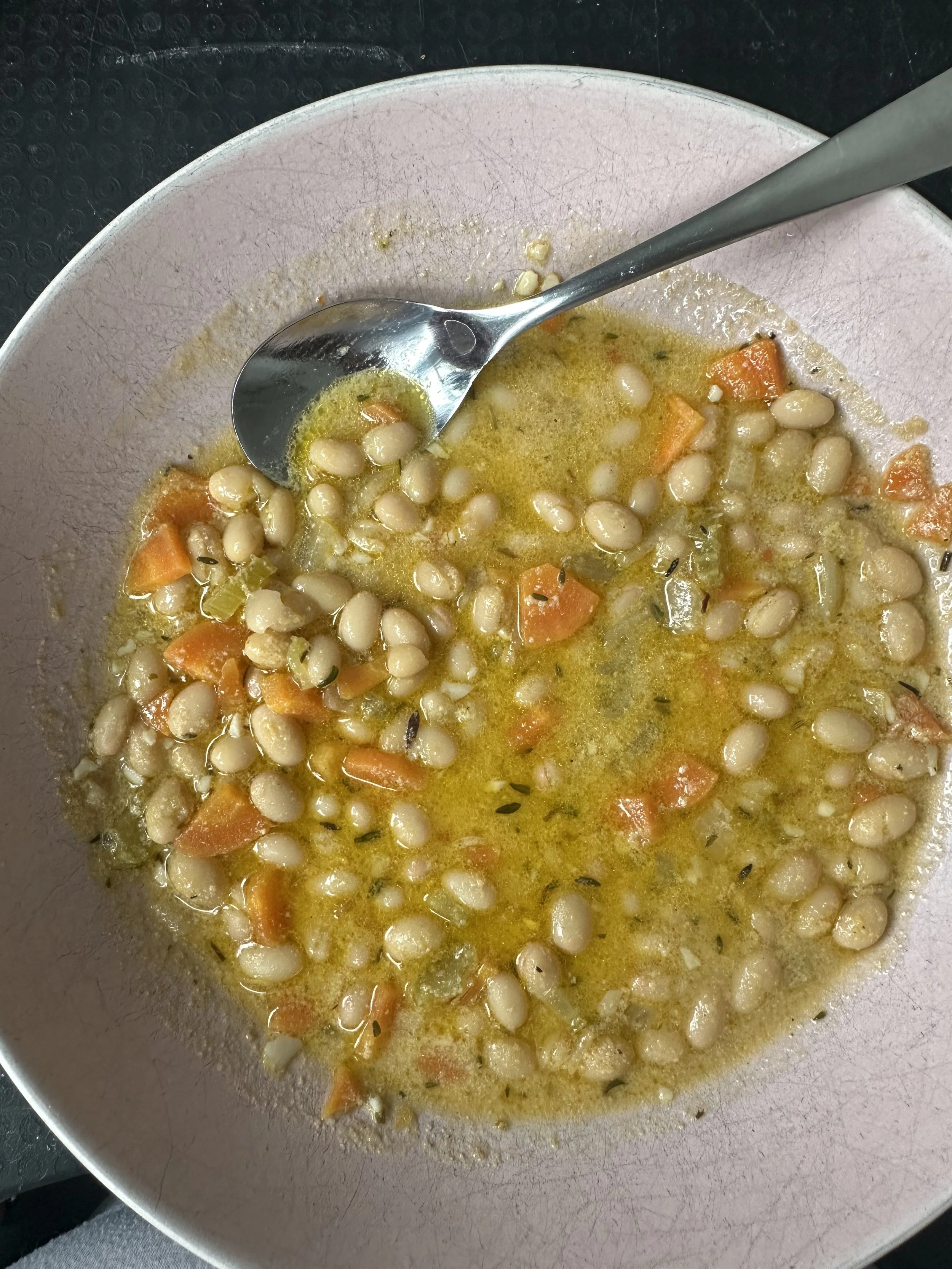 A bowl of white bean and vegetable soup with carrots, herbs, and broth, with a spoon inside.