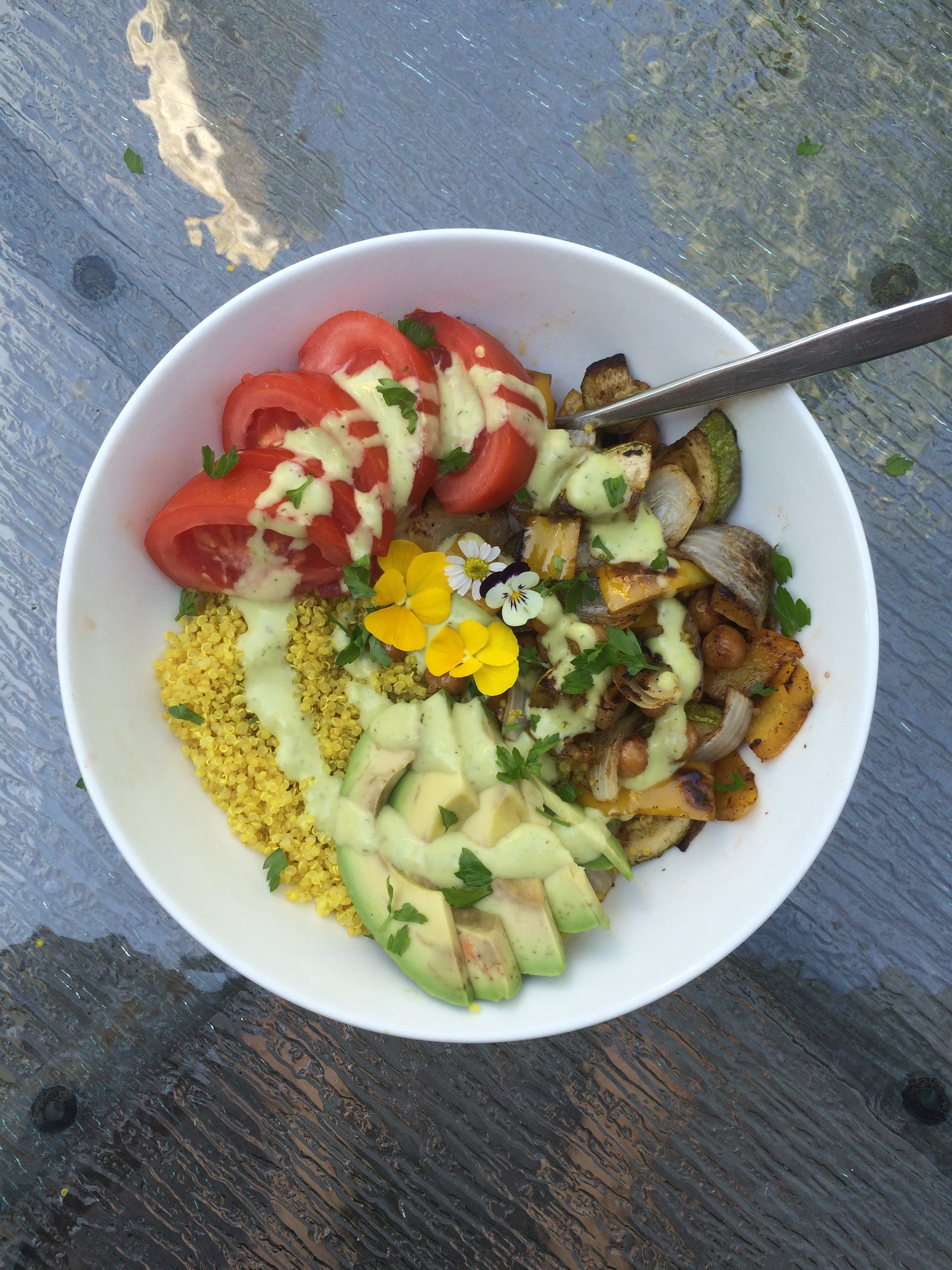 A bowl of food with sliced tomatoes, sliced avocado, cooked vegetables topped with green sauce, and garnished with edible flowers and chopped herbs, placed on a textured surface.